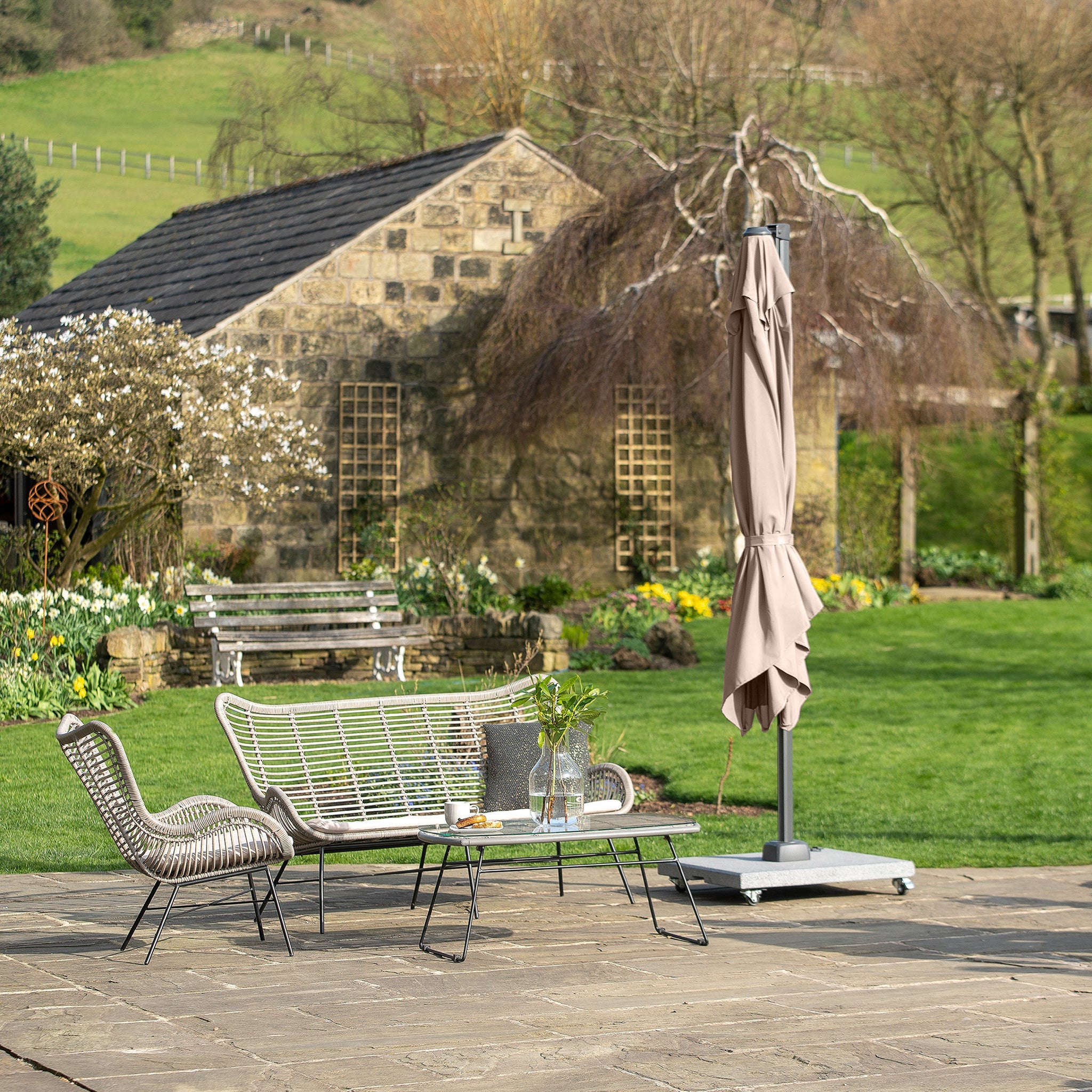 A patio featuring wicker furniture and a Pacific Lifestyle - Voyager T2 2.7m Square Parasol in Grey, offering UPF35+ protection, is set against a stone building with lush greenery. A closed umbrella stands nearby.