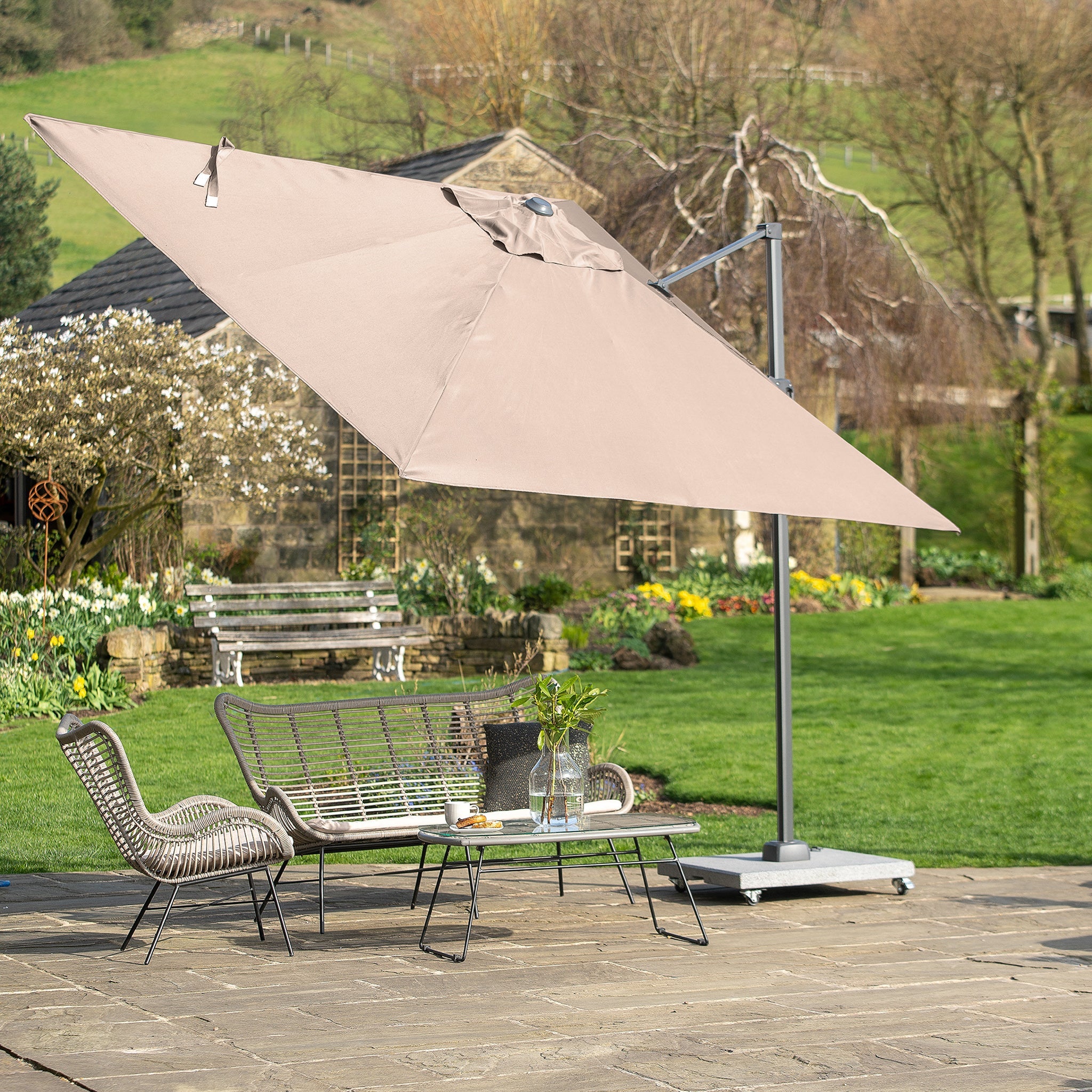 Outdoor patio featuring a Pacific Lifestyle Voyager T2 2.7m Square Parasol in Grey with UPF35+ protection, alongside wicker furniture and a serene garden view.