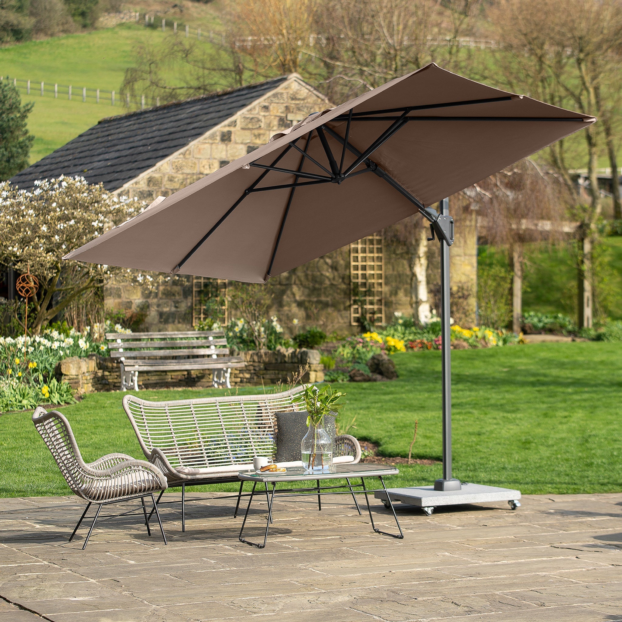 A patio set with a wicker sofa, chairs, and a Pacific Lifestyle Voyager T2 2.7m Square Parasol in Grey on a stone surface provides UPF35+ protection. A charming garden and brick building complete the backdrop.