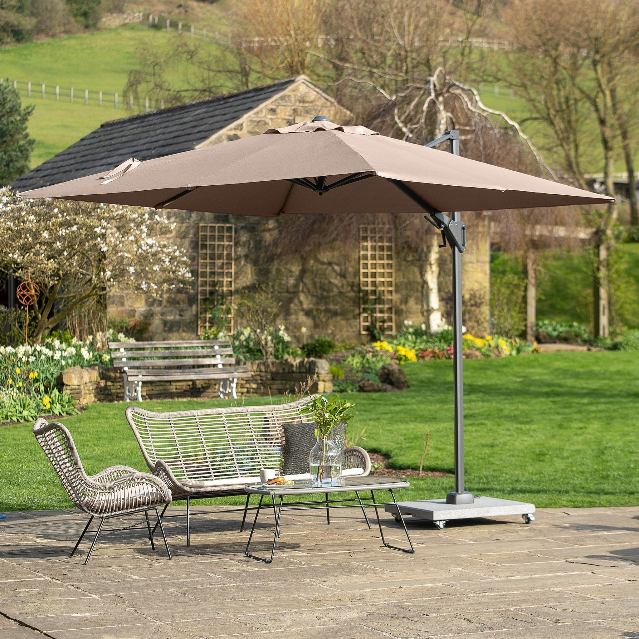Patio scene with a large Voyager T2 2.7m Square Parasol in Grey featuring an aluminium pole and 360° tilt, casting shade over a metal bench, two chairs, a coffee table, and lush green grass in the background.