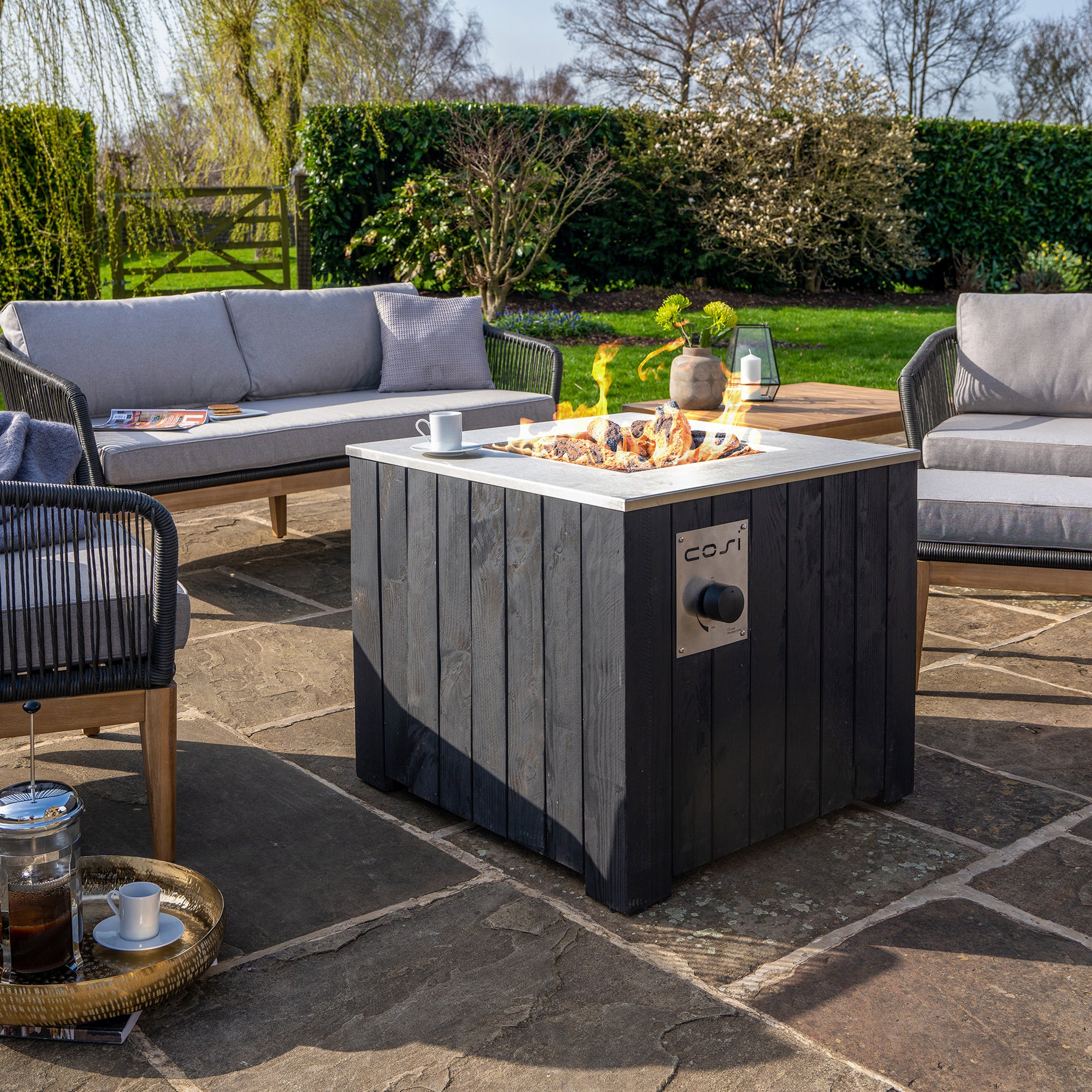 Outdoor patio setting featuring a Cosi - Cosicube 70 Black Fire Pit, surrounded by gray sofas and lush greenery in the background.