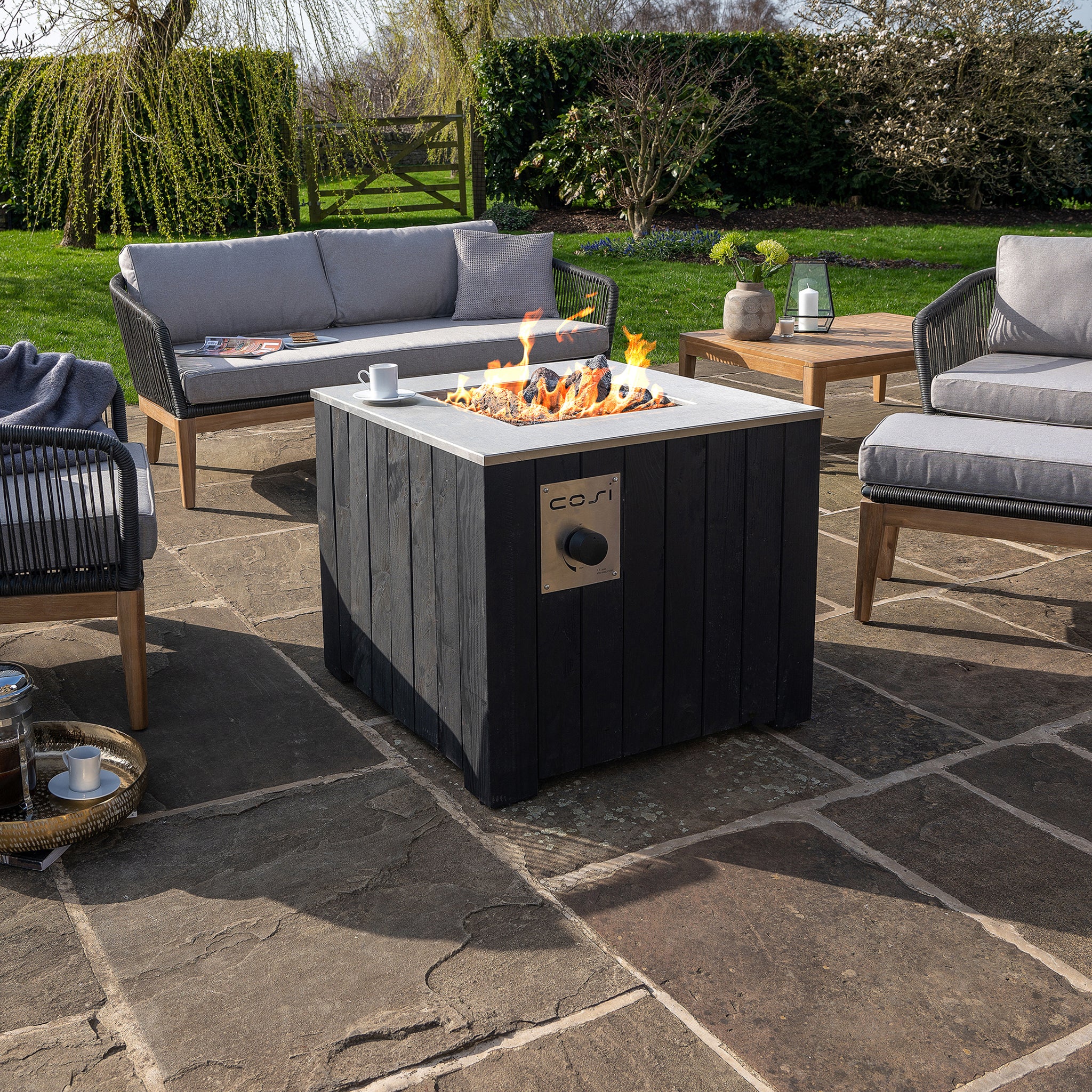Outdoor patio featuring a Cosi - Cosicube 70 Black Fire Pit with gray sofas and a wooden table on stone paving, using clean propane gas for seamless warmth and ambiance.