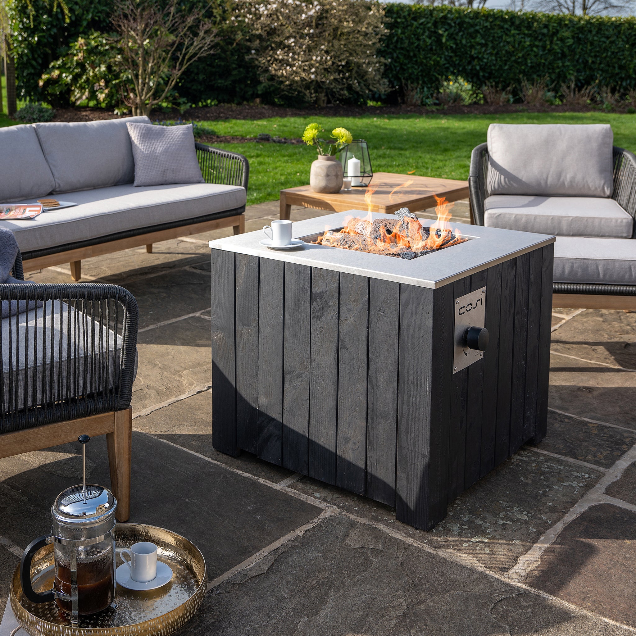 Outdoor patio with wicker furniture centered around a Cosi - Cosicube 70 Black Fire Pit table. Coffee and cups rest invitingly on a tray in the foreground.