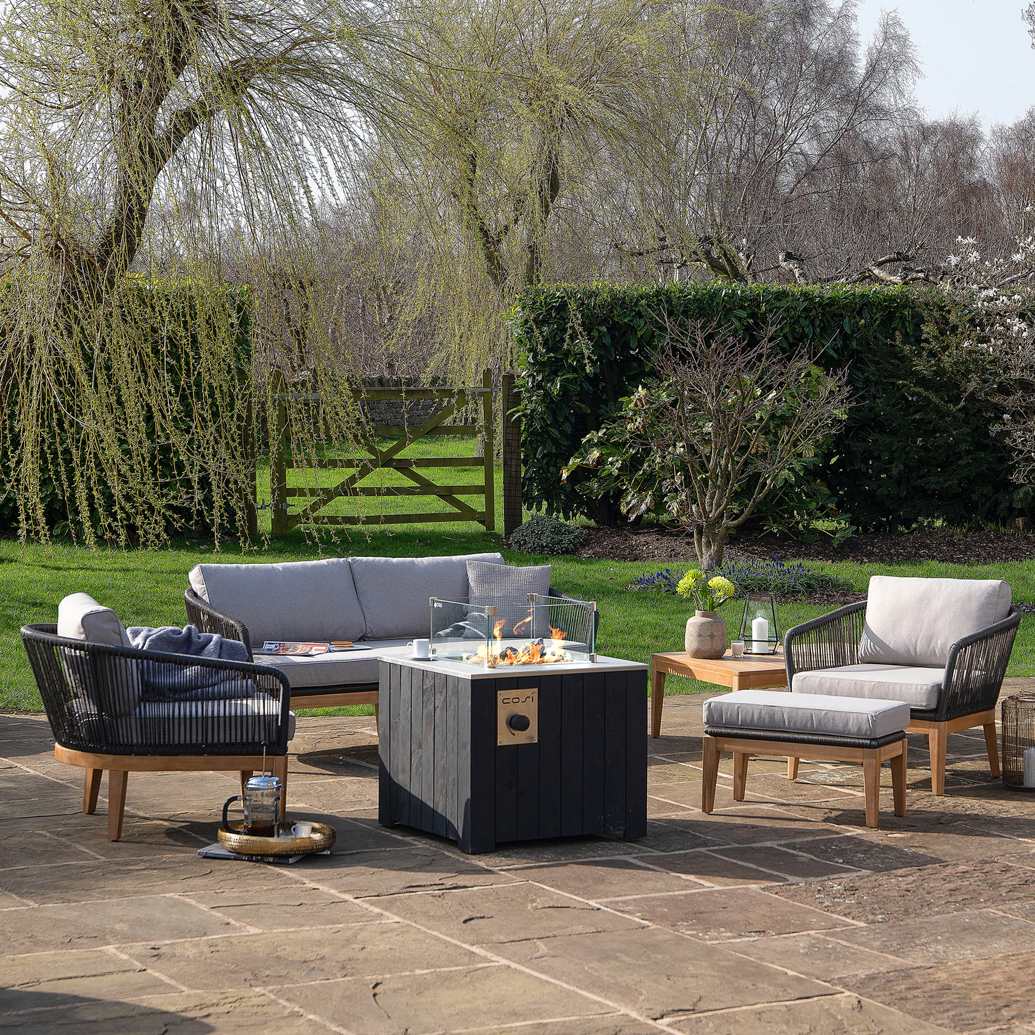 An outdoor patio features a cozy sofa and chairs centered around the Cosi - Cosicube 70 Black Fire Pit on stone paving, surrounded by lush greenery and trees.
