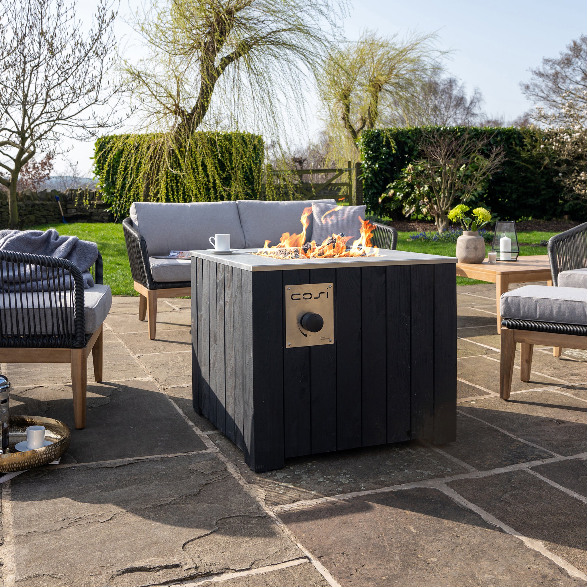 The outdoor patio features a Cosi - Cosicube 70 Black Fire Pit, fueled by propane gas, surrounded by cushioned chairs, plants, and trees.
