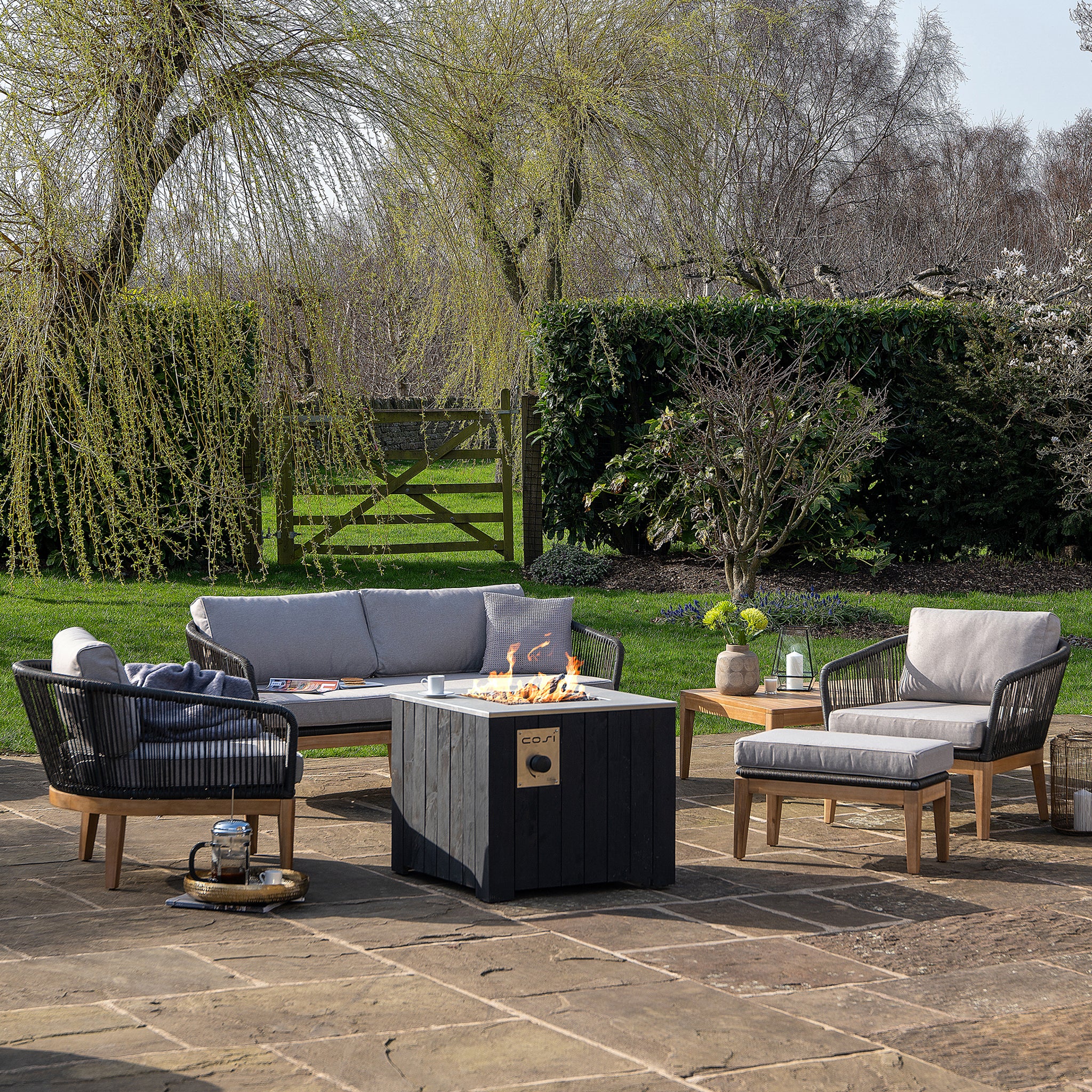 The outdoor patio showcases the Cosi - Cosicube 70 Black Fire Pit, surrounded by a sofa, chairs, and side tables. Amid trees and greenery on a sunny day, it creates the ideal propane-powered space for relaxation or entertaining.