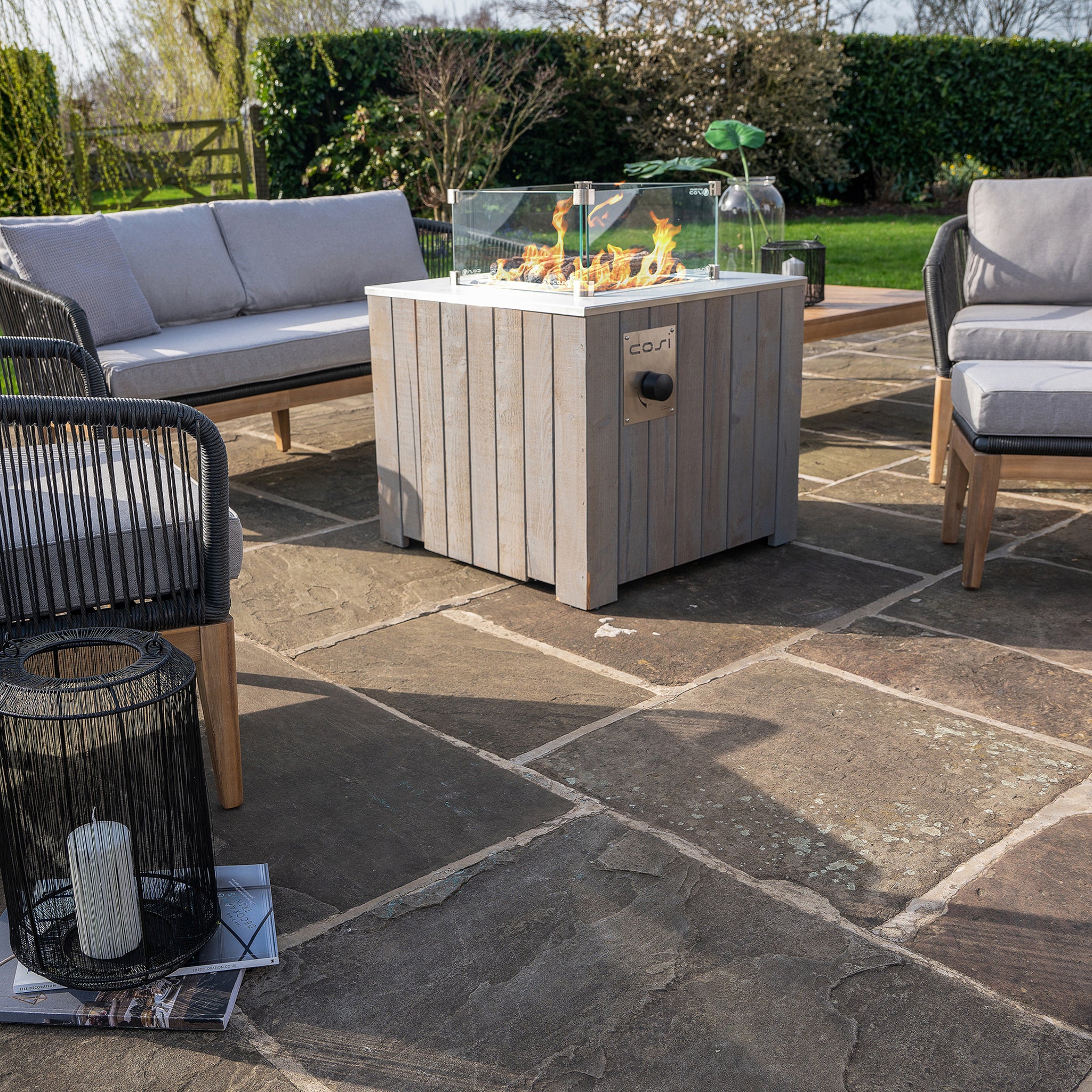Outdoor patio with a Cosi Cosicube 70 Grey Wash Fire Pit, grey chairs, and lantern on stone tiles, set in a lush garden.