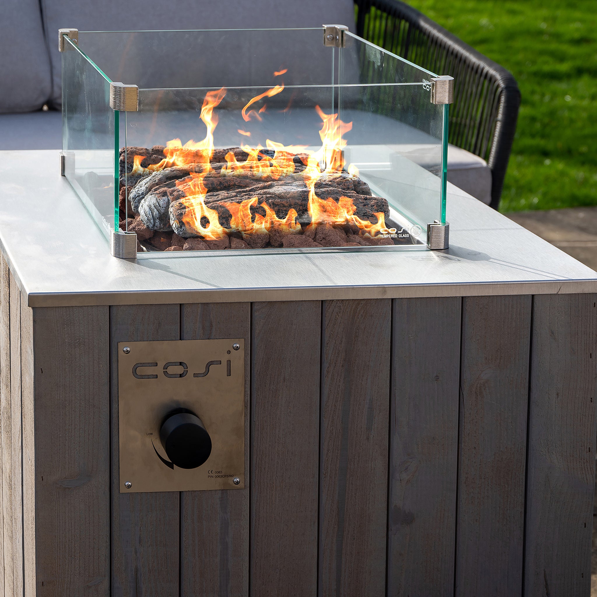 The Cosi Cosicube 70 Grey Wash Fire Pit is a modern outdoor fire pit with glass walls and a wooden base, showcasing beautiful flames over simulated logs, all efficiently powered by propane gas.