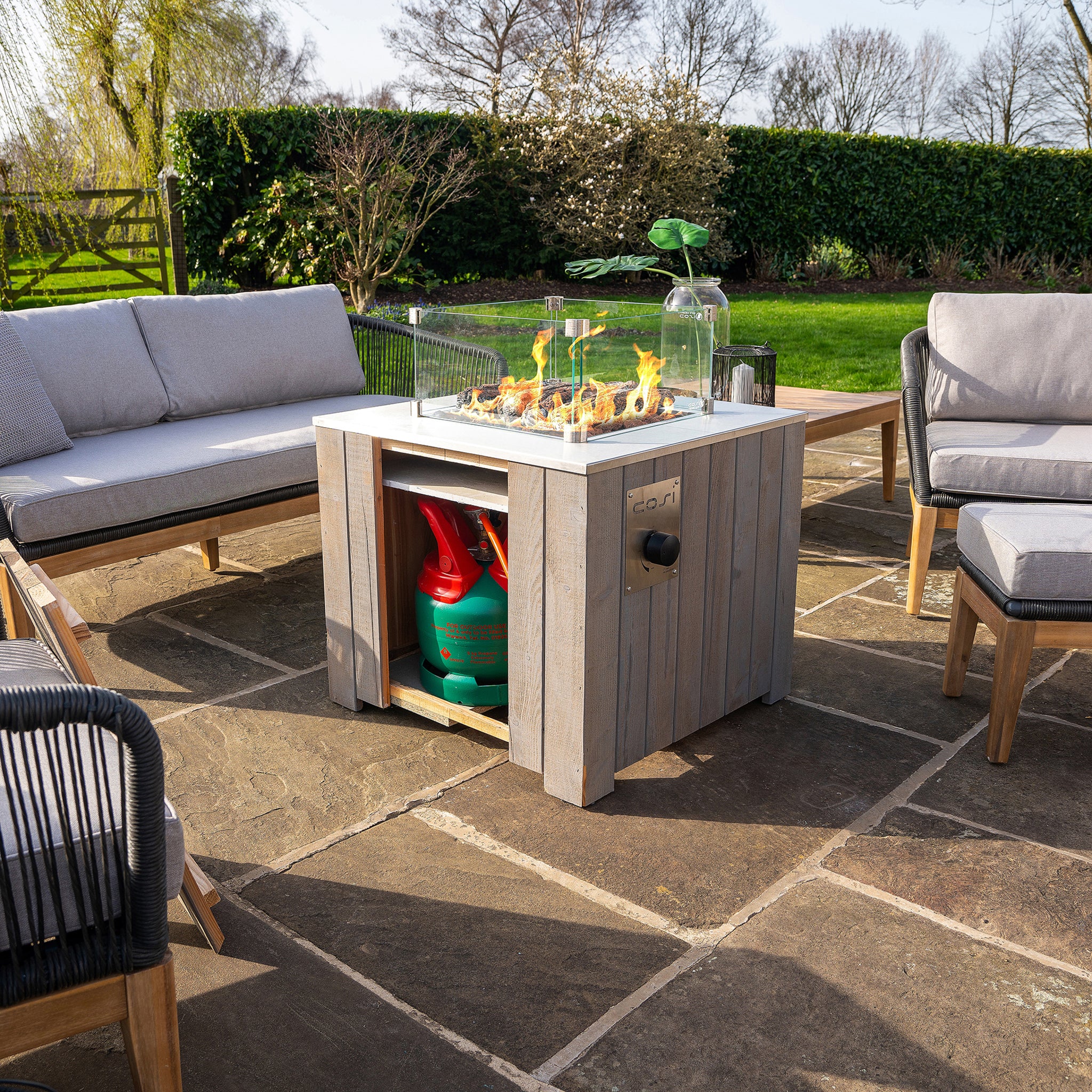 An outdoor patio features gray couches and a wooden Cosi - Cosicube 70 Grey Wash Fire Pit table on a stone floor, surrounded by lush greenery.