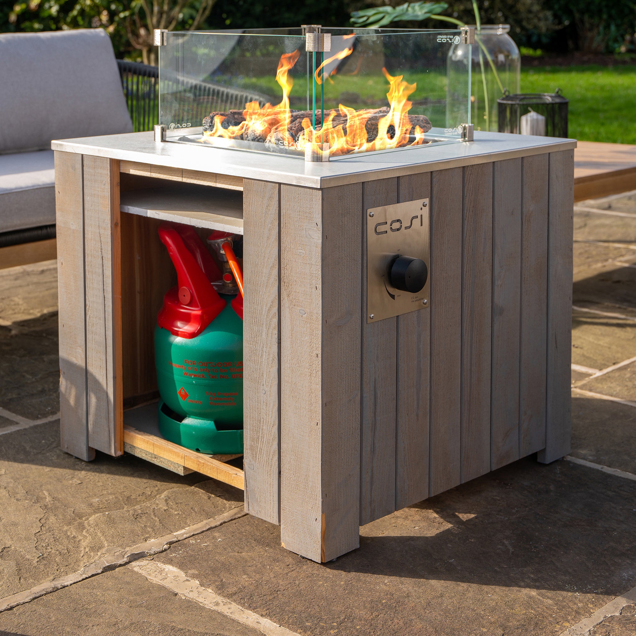 The Cosi Cosicube 70 Grey Wash Fire Pit includes a glass guard and displays a visible propane tank beneath, perfectly set on a stone patio.