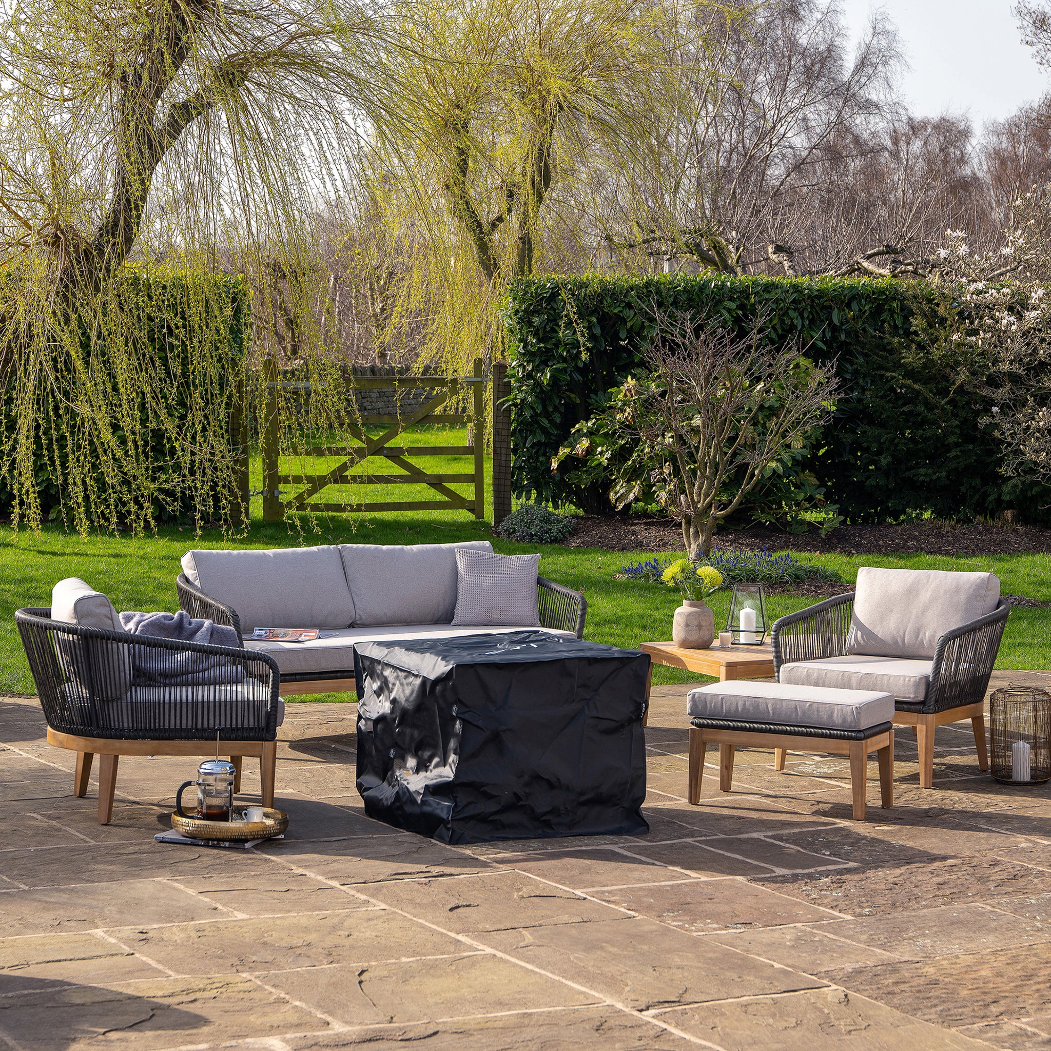 An outdoor patio showcases a chic wicker furniture set encircling a Cosi - Cosicube 70 Grey Wash Fire Pit, amidst lush greenery and towering trees.