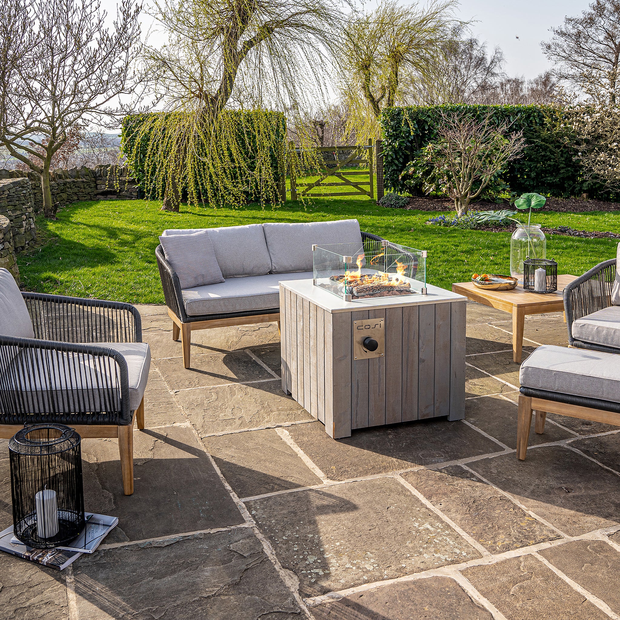 A patio features a comfortable seating area and the Cosi - Cosicube 70 Grey Wash Fire Pit, fueled by propane, set on stone tiles and surrounded by lush grass and trees.