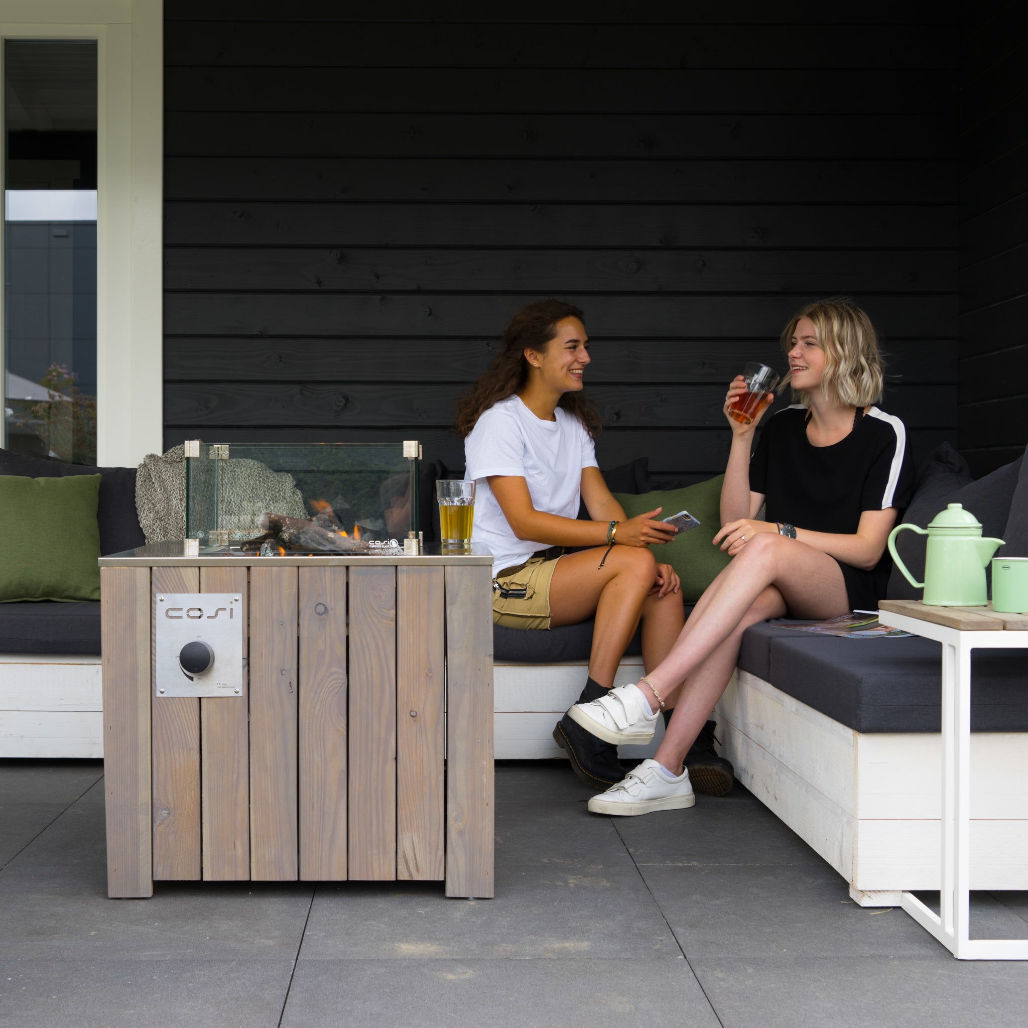 Two people relax on a patio couch, chatting and enjoying drinks beside the warm glow of a Cosi - Cosicube 70 Grey Wash Fire Pit, powered by clean-burning propane gas.