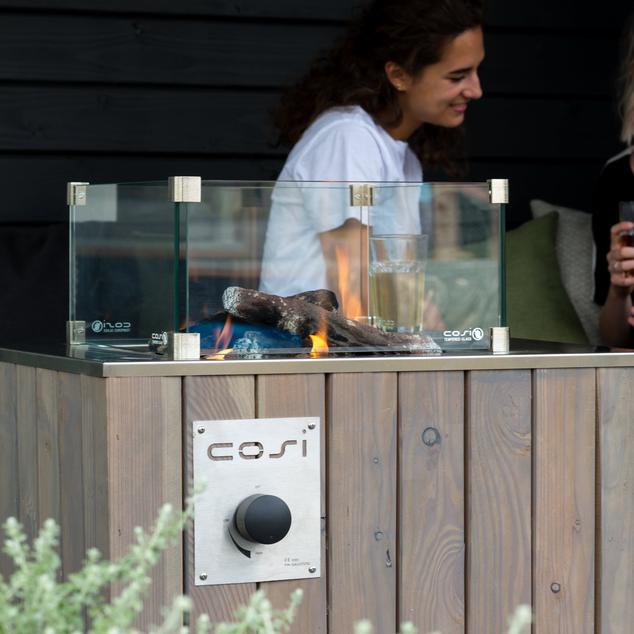 Two individuals sit beside a stylish Cosi - Cosicube 70 Grey Wash Fire Pit, their wooden table featuring a glass-enclosed outdoor fire pit.