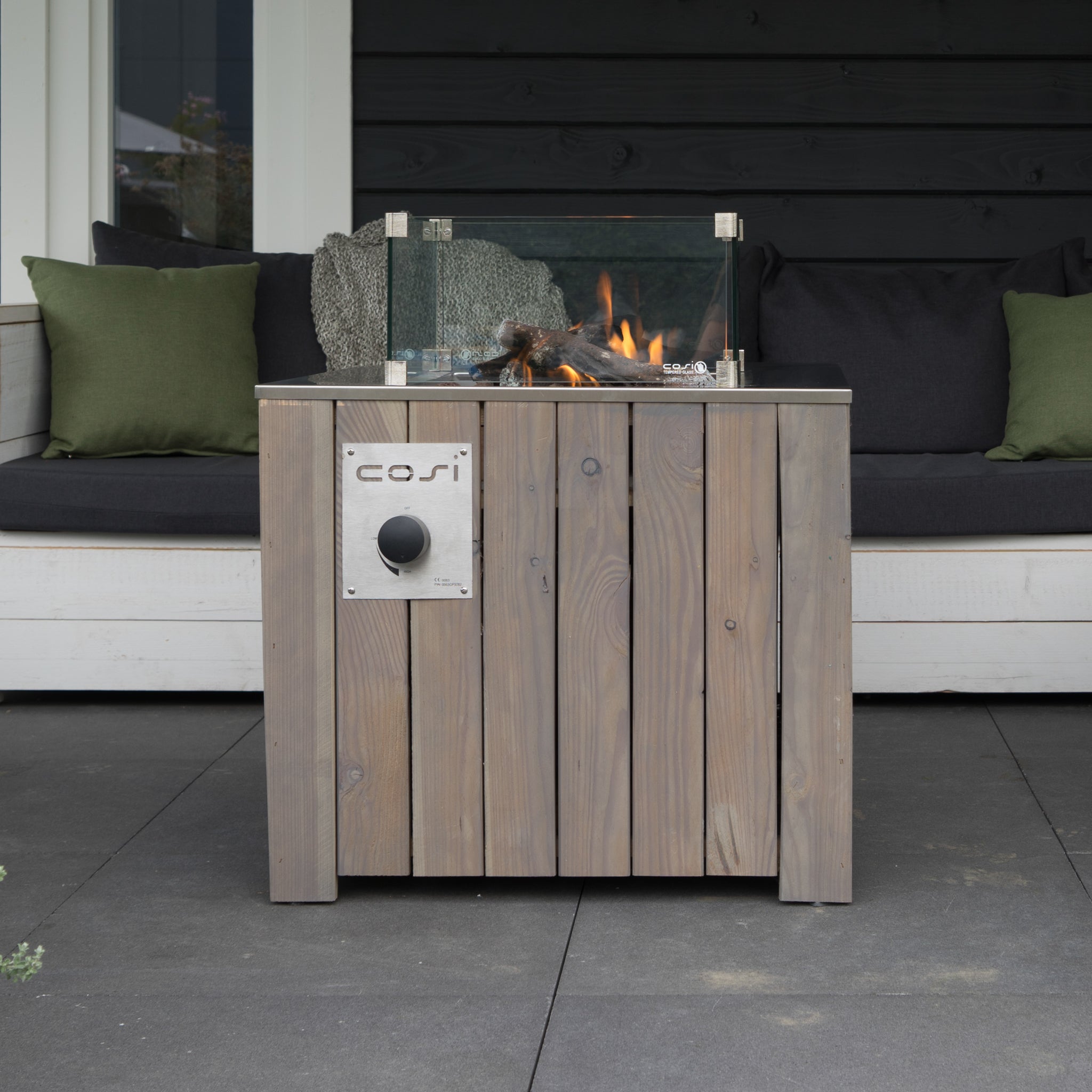 The Cosi - Cosicube 70 Grey Wash Fire Pit, equipped with a glass windguard, enhances the patio's ambiance. It's encircled by a white cushioned sofa accented with green pillows.