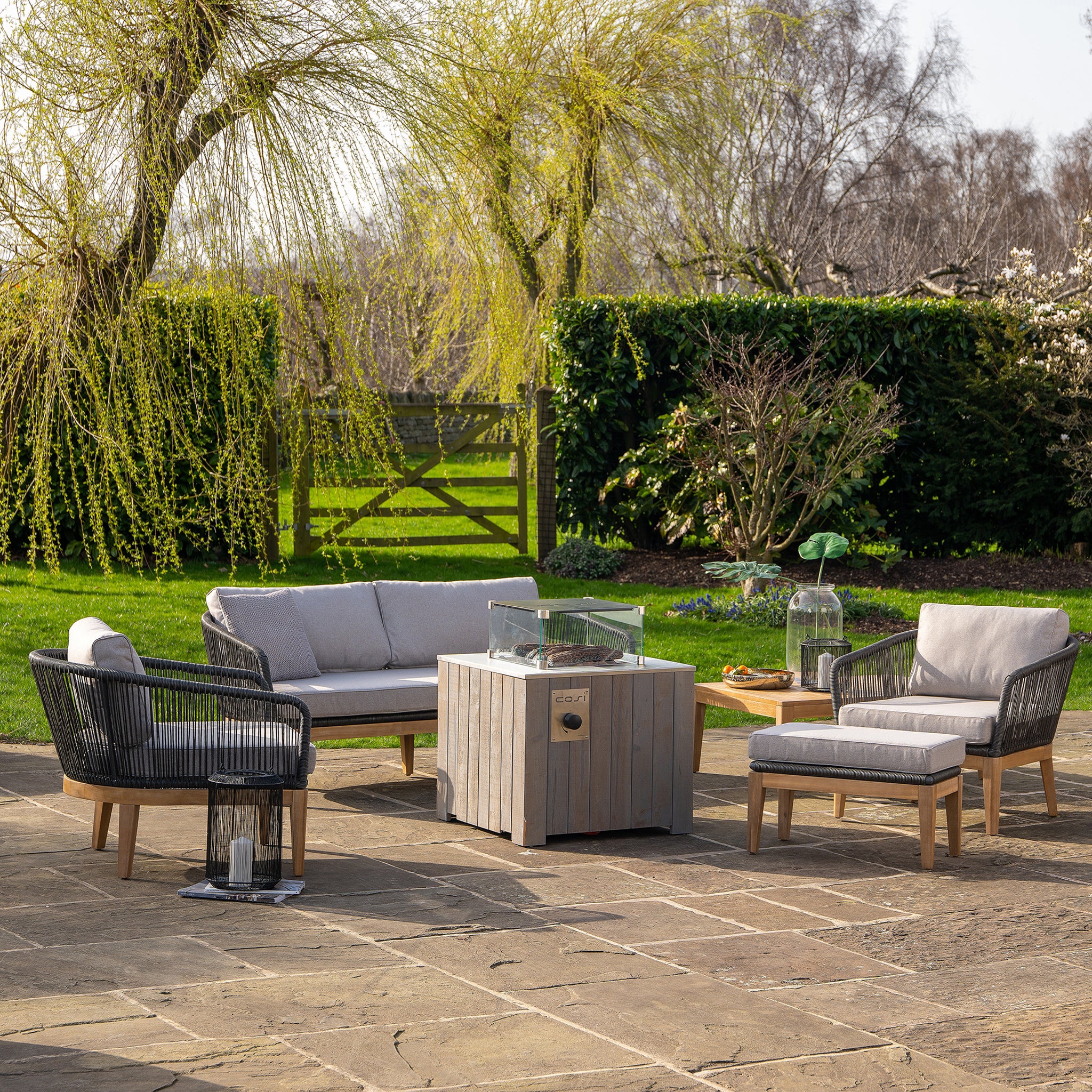 A cozy patio with wicker furniture surrounds a Cosi - Cosicube 70 Grey Wash Fire Pit, nestled among trees and lush greenery under a clear sky.