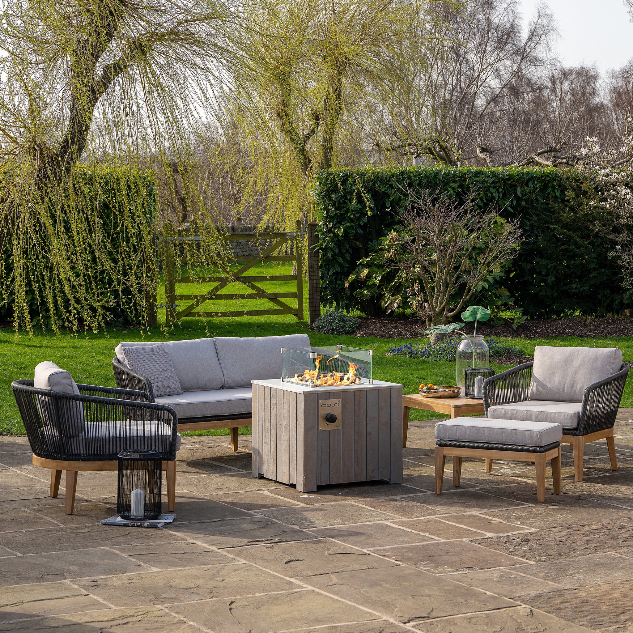 Outdoor patio featuring modern furniture and the Cosi - Cosicube 70 Grey Wash Fire Pit table, set against a lush garden backdrop.