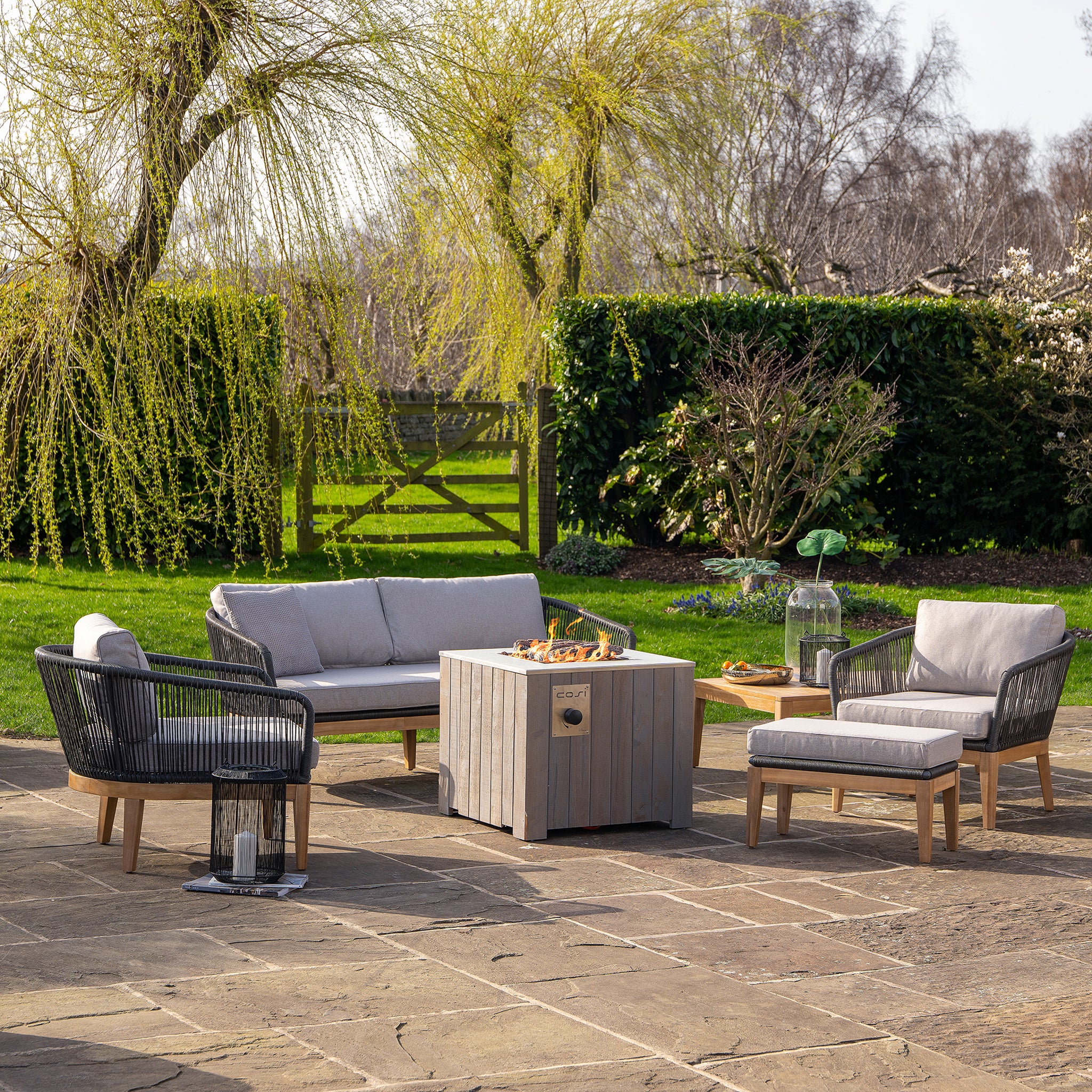 A trendy outdoor patio set with gray cushions features the Cosi Cosicube 70 Grey Wash Fire Pit, nestled among vibrant greenery beneath a clear sky.