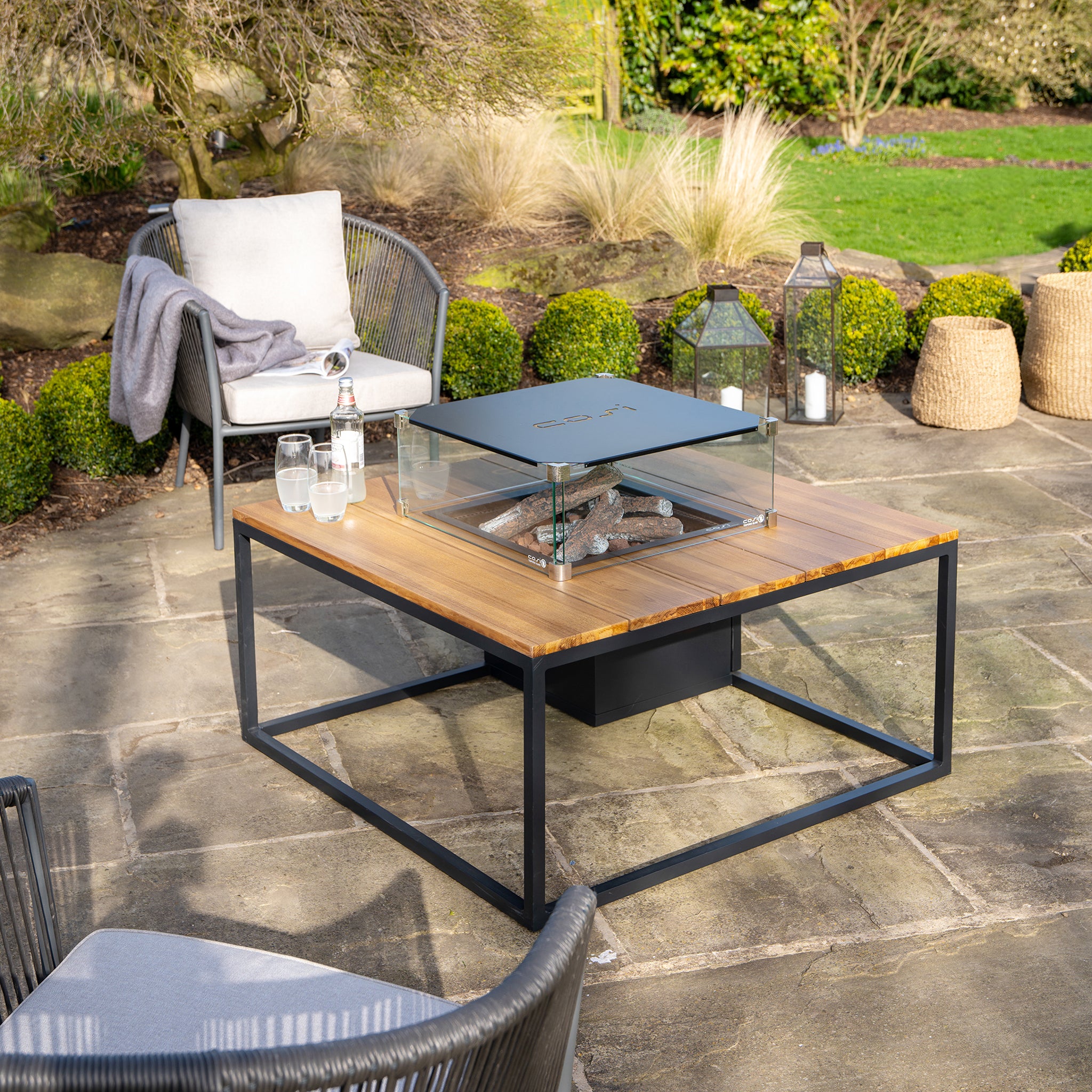 Outdoor patio highlights the Cosi Cosiloft 100 Black and Teak Fire Pit Table, surrounded by chairs and lush greenery.