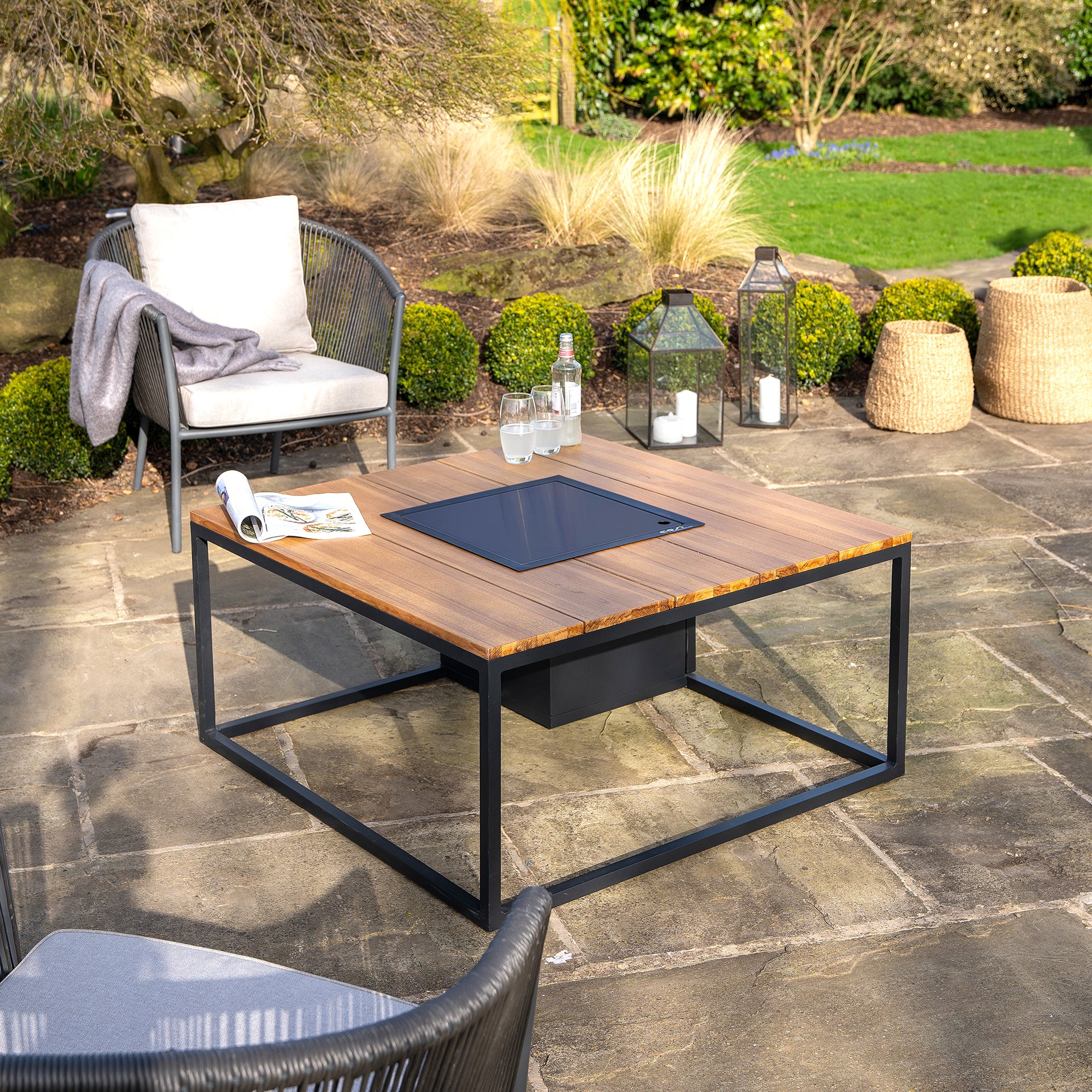 An inviting outdoor patio features a Cosi Cosiloft 100 Black and Teak Fire Pit Table adorned with water bottles, a magazine, and two chairs. Nestled in a lush garden setting, this weatherproof ensemble promises durability and style throughout the seasons.