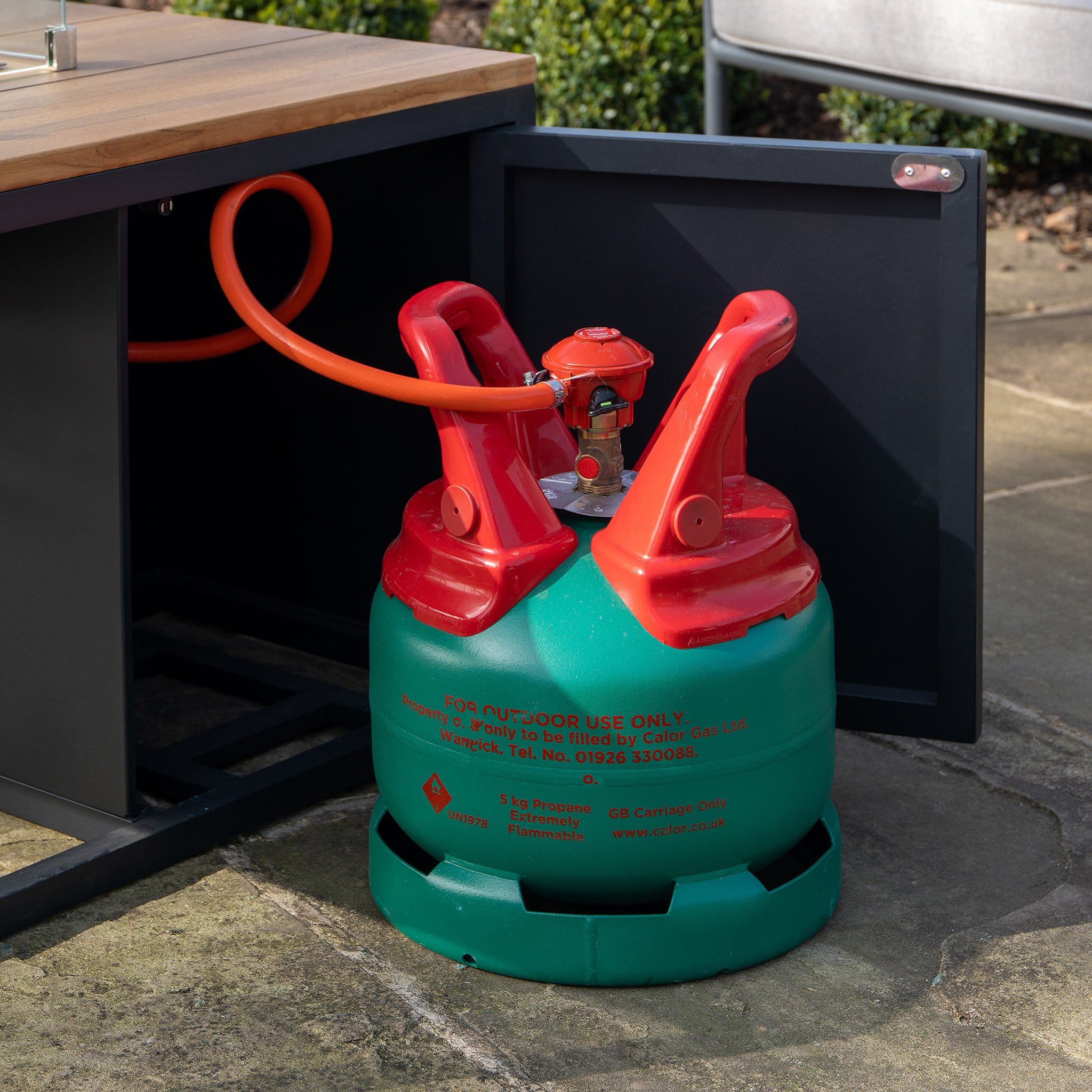 A Cosi - Cosiloft 100 Black and Teak Fire Pit Table featuring a red and green gas cylinder, connected by an orange hose, is partially nestled inside.