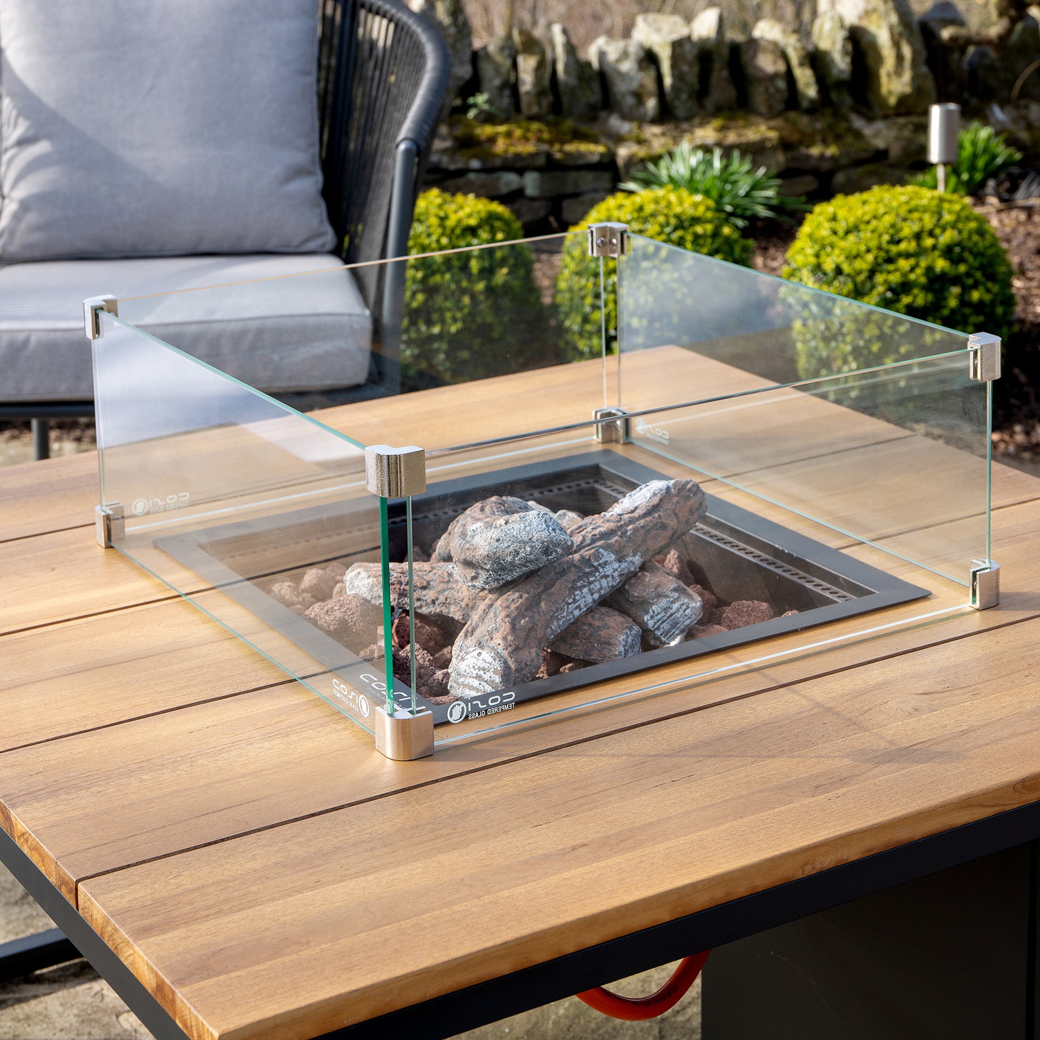The Cosi Cosiloft 100 Black and Teak Fire Pit Table, featuring decorative stones and a glass enclosure, elegantly enhances any setting with its weatherproof design.