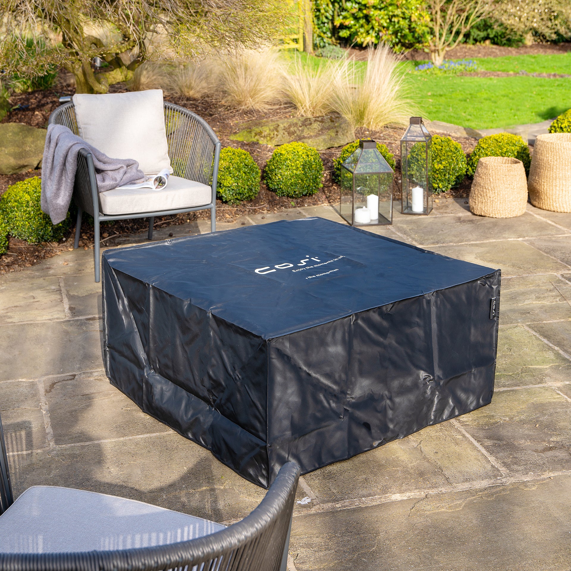 A Cosi - Cosiloft 100 Black and Teak fire pit table on the patio is surrounded by weatherproof wicker chairs and lush greenery.
