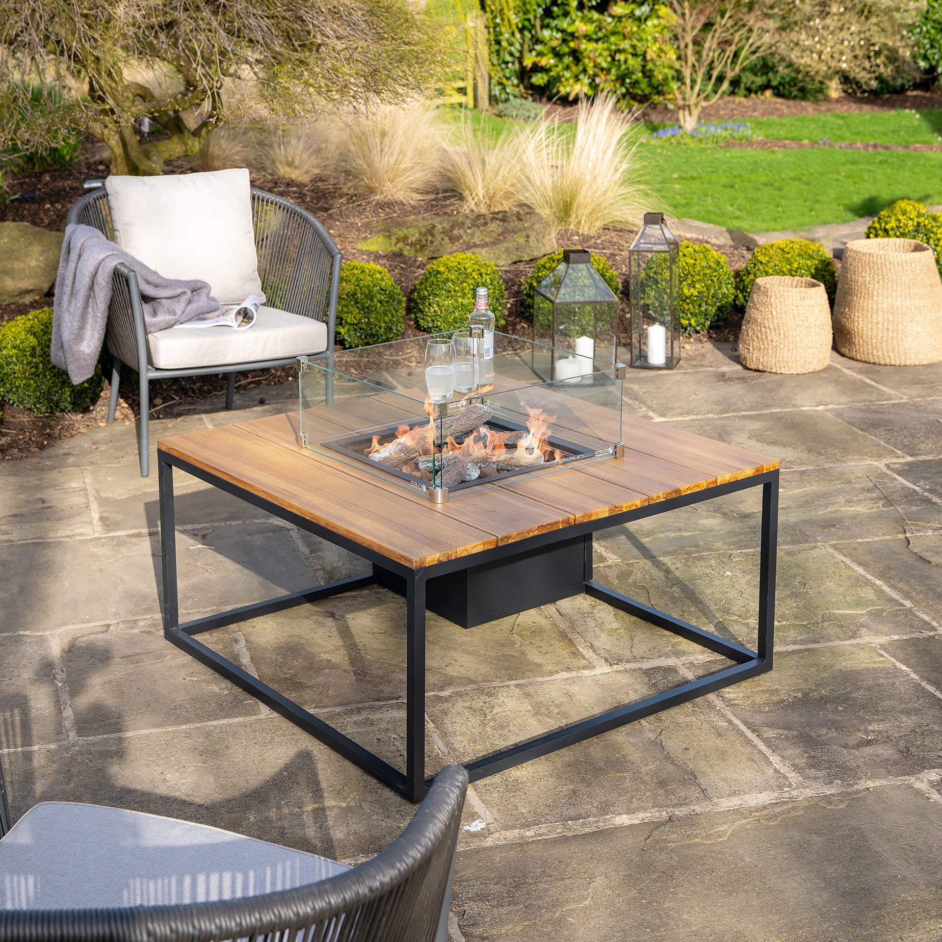 A patio showcasing the Cosi Cosiloft 100 Black and Teak Fire Pit Table, enhanced by teak wood seating and lanterns, set against lush garden greenery.