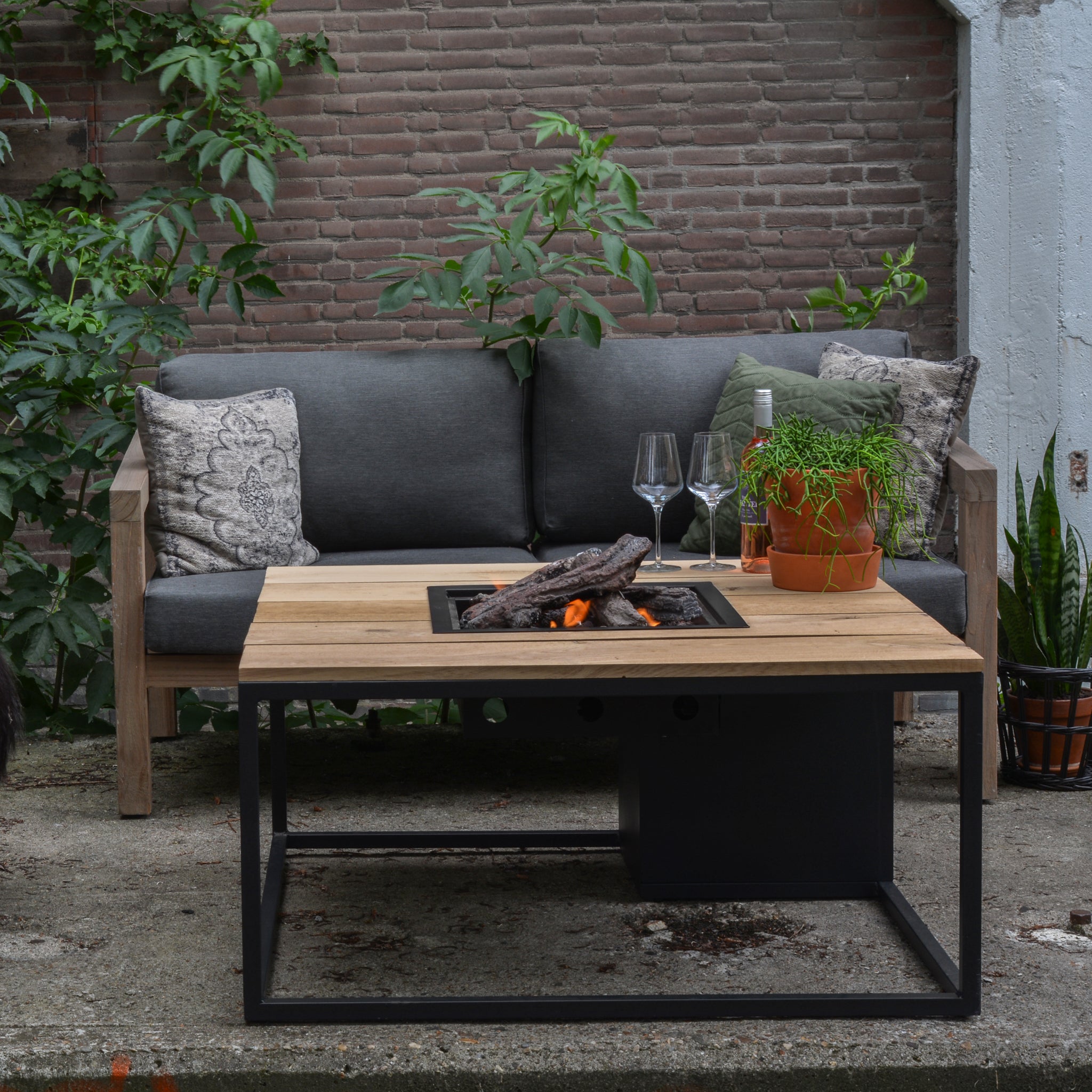 Outdoor seating area with the Cosi - Cosiloft 100 Black and Teak Fire Pit Table, a gray sofa, lush plants, and wine glasses on a weatherproof brick patio.