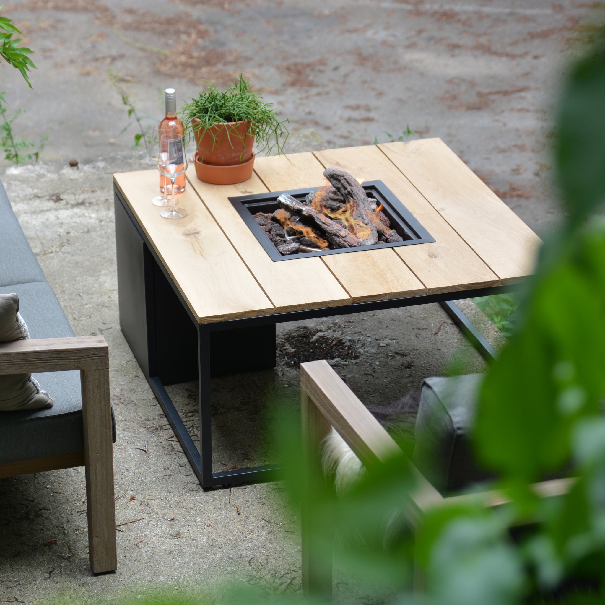 The Cosi - Cosiloft 100 Black and Teak Fire Pit Table, weatherproof and featuring a built-in pit, is surrounded by chairs, a plant, and a bottle of rosé wine.
