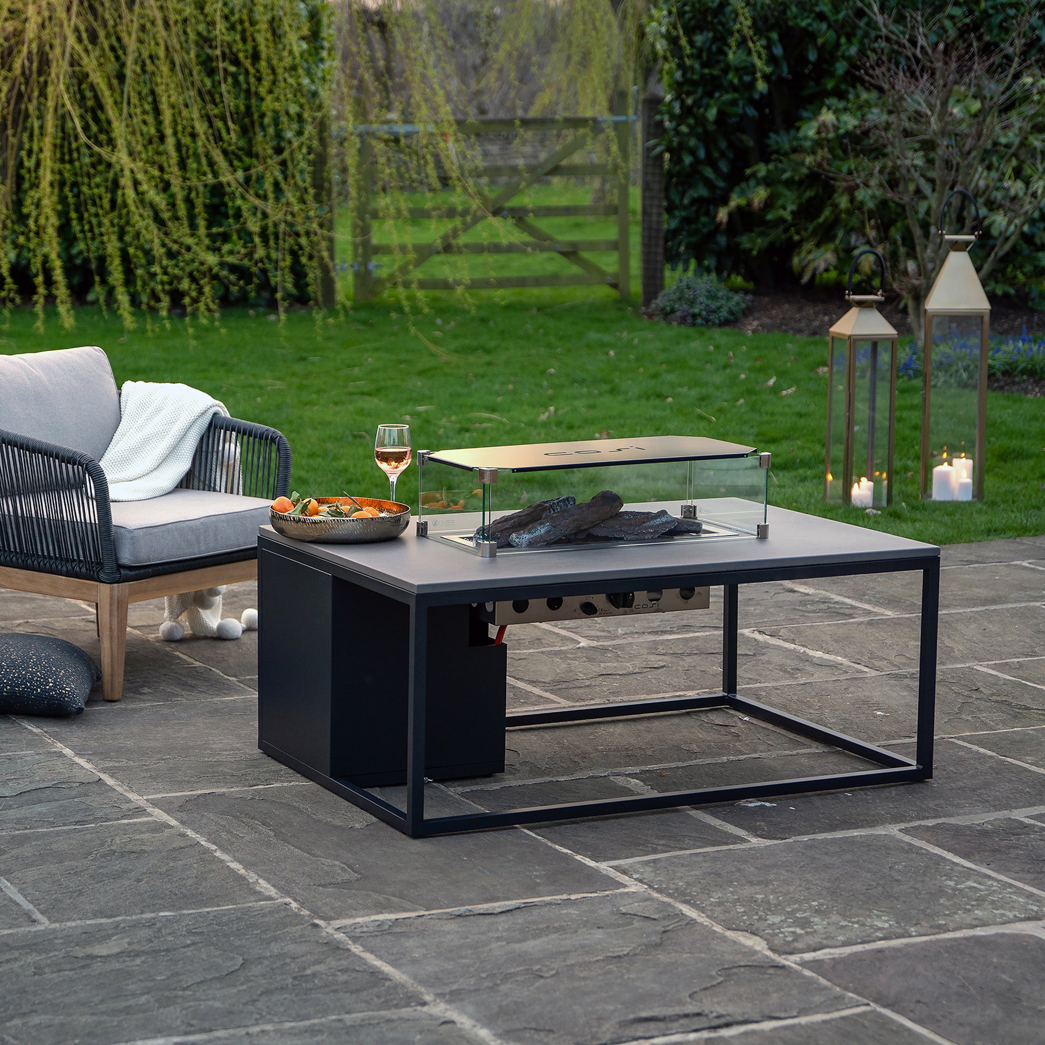 An elegant outdoor lounge boasts the Cosi Cosiloft 120 Black and Grey Fire Pit Table, ideal for relaxing with drinks and snacks. Cozy chairs and lanterns enhance the warm ambiance in a lush garden setting, seamlessly elevating style and comfort.