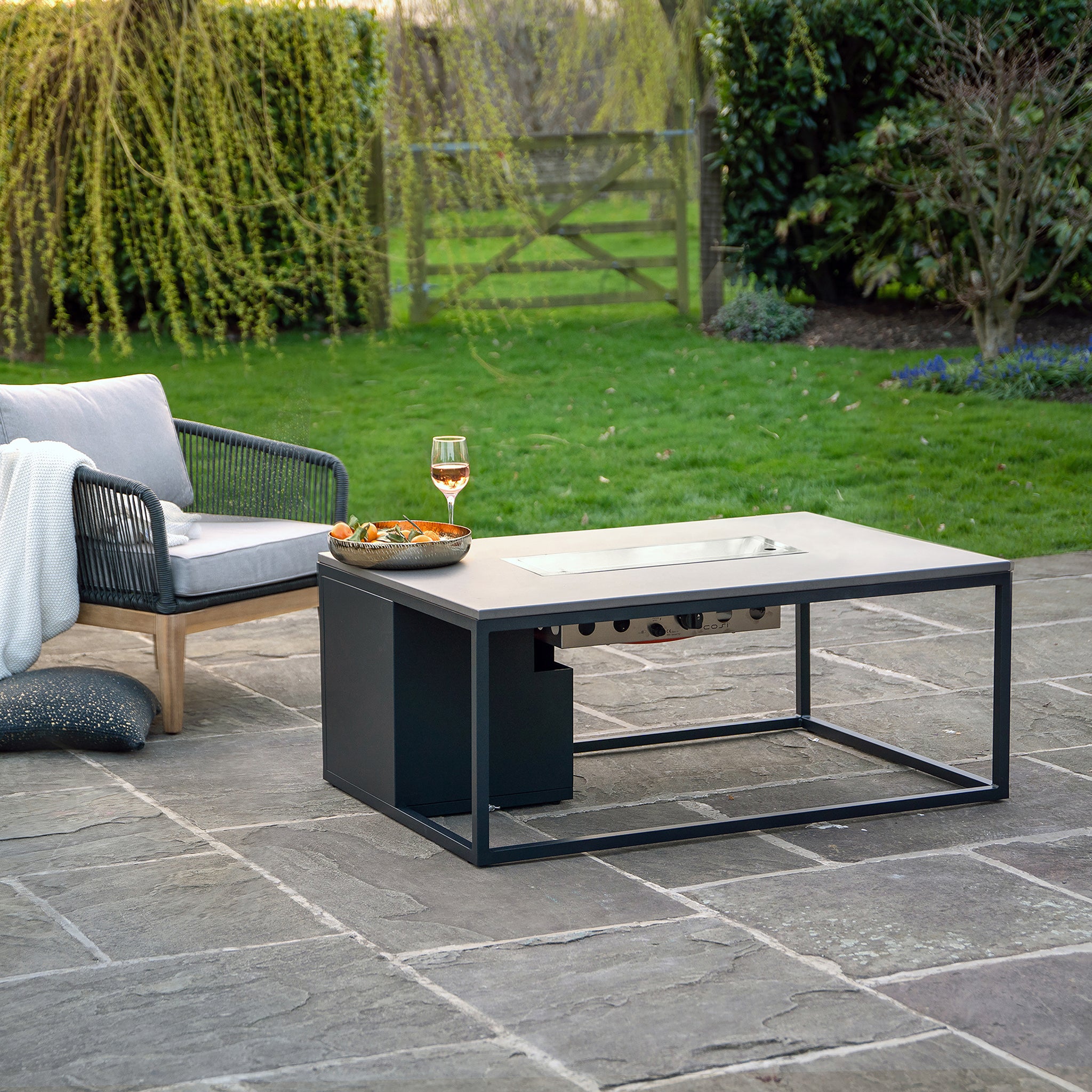 An outdoor lounging area showcases a Cosi - Cosiloft 120 Black and Grey Fire Pit Table with snacks and wine, next to a cushioned chair on stone pavement.
