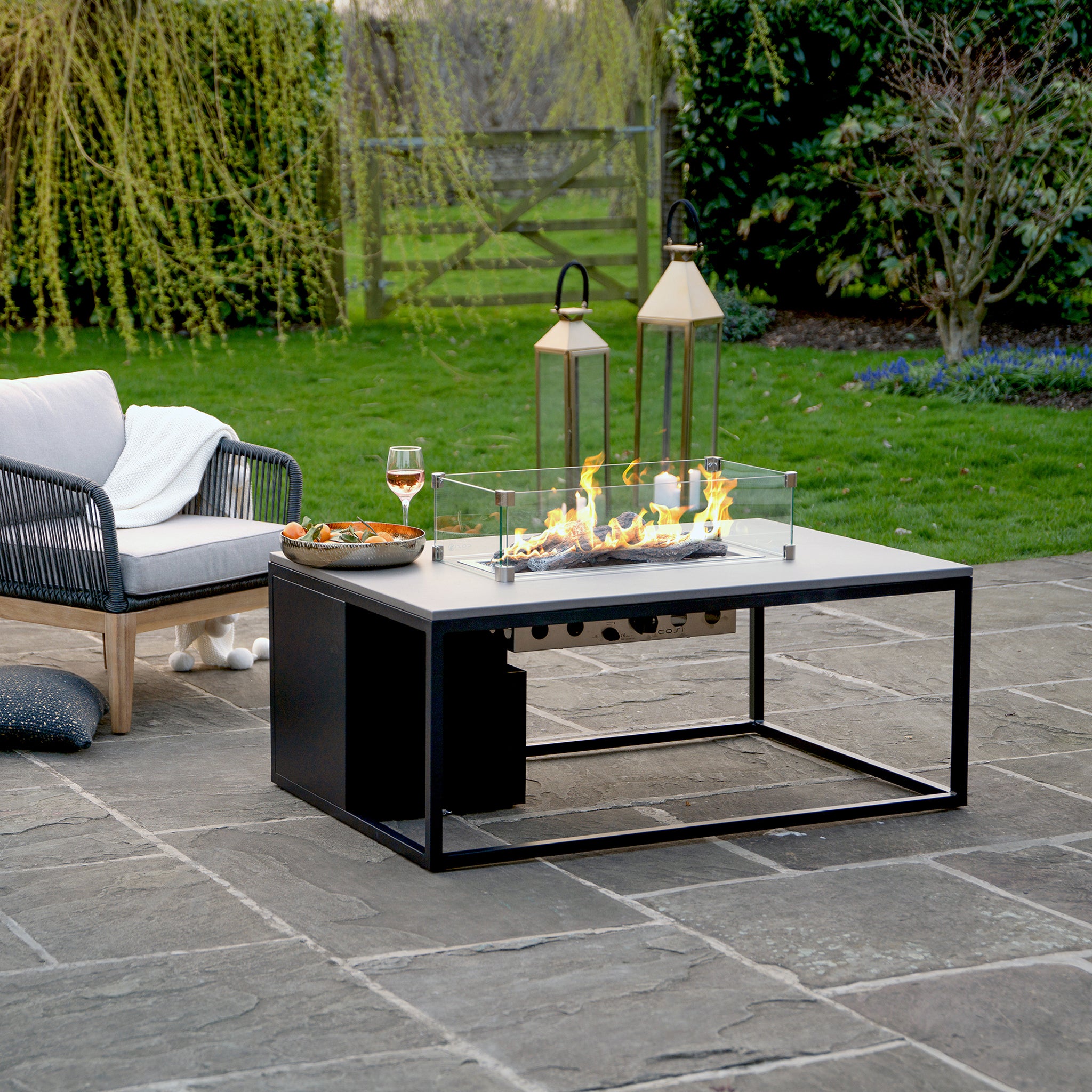 A patio with a Cosi Cosiloft 120 Black and Grey Fire Pit Table, wine glass, snacks, lanterns, and cushioned chair on stone tiles offers a cozy outdoor lounging area.