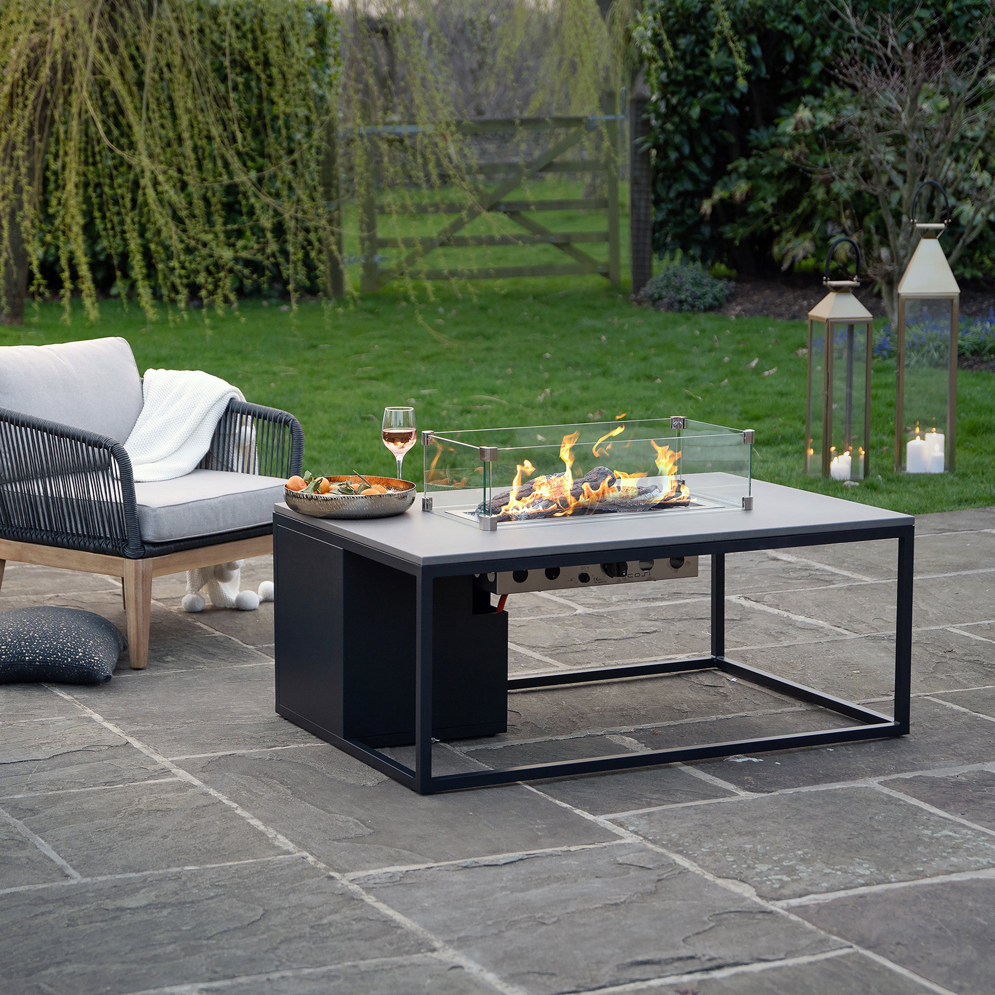 An outdoor patio showcases a Cosi Cosiloft 120 Black and Grey Fire Pit Table, alongside a cushioned chair and lanterns on stone. This setup creates the ideal lounging spot, complemented by greenery in the background.
