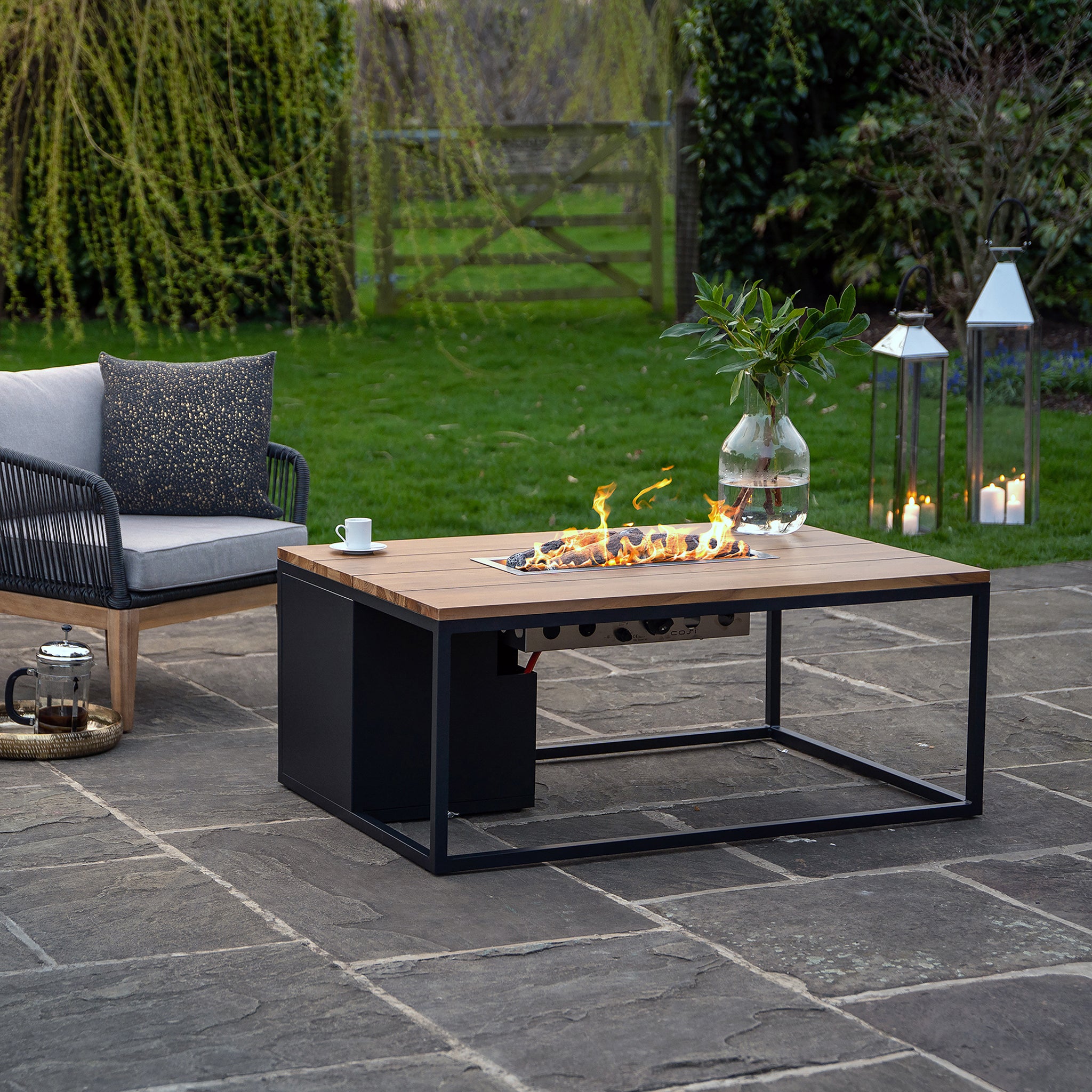 An outdoor patio with a teak couch, Cosi Cosiloft 120 Black and Teak Fire Pit Table with a concrete effect, lanterns, and lush greenery.