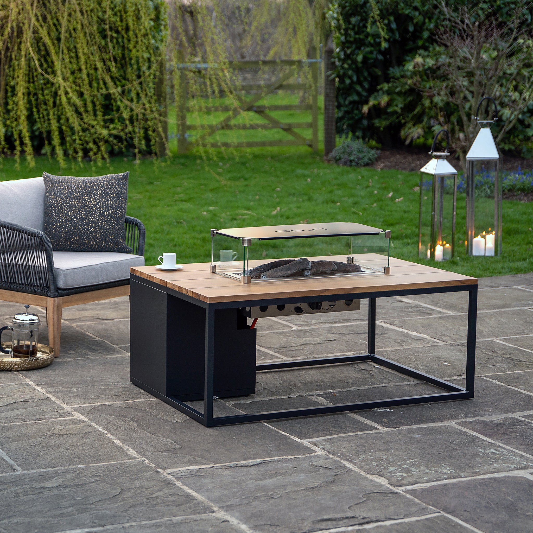 On the stone patio, the Cosi - Cosiloft 120 Black and Teak Fire Pit Table stands surrounded by a teak wood sofa and lanterns, creating a modern look against the lush green garden backdrop.