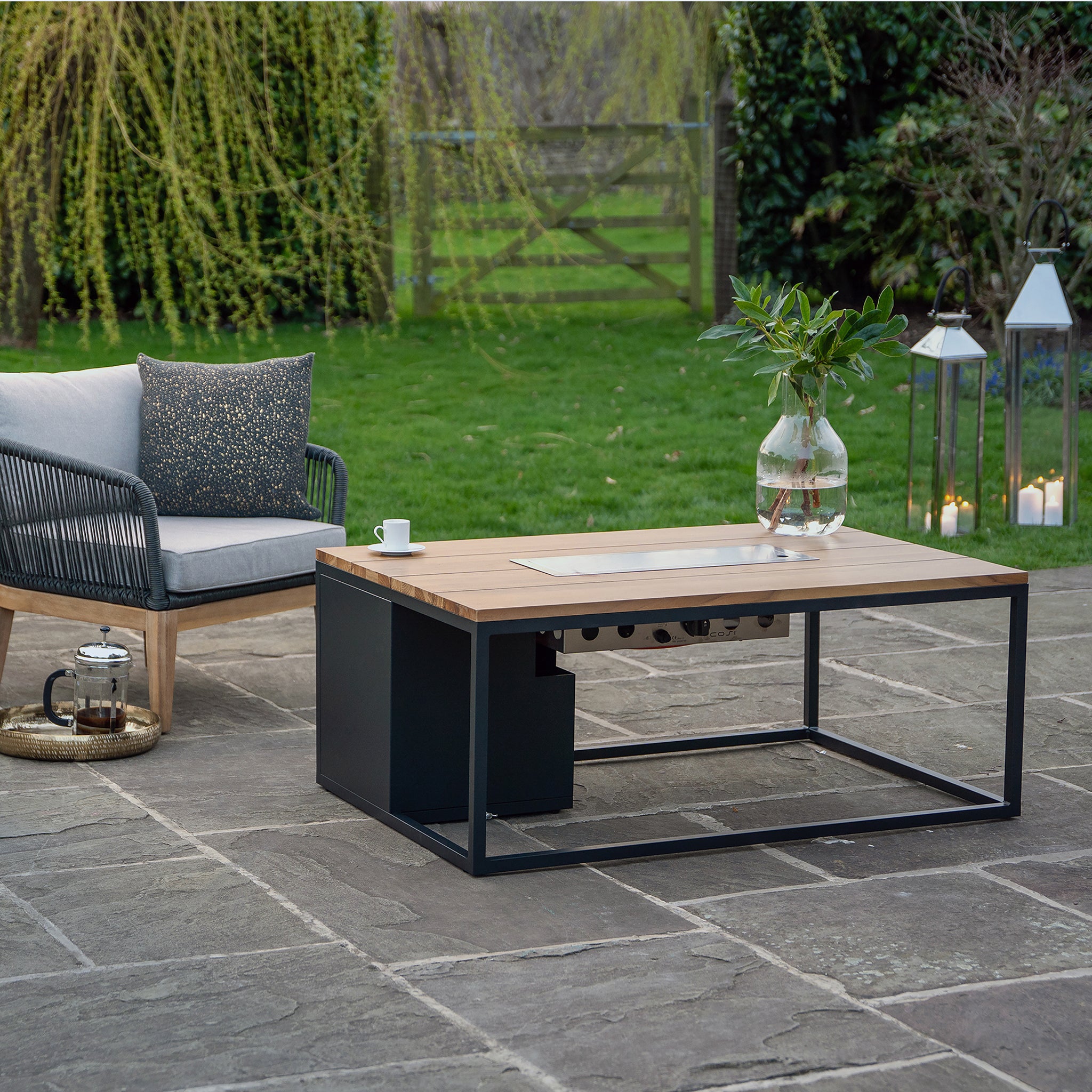 Outdoor seating features the Cosi - Cosiloft 120 Black and Teak Fire Pit Table on a stone patio; candles and plants create a serene ambiance.