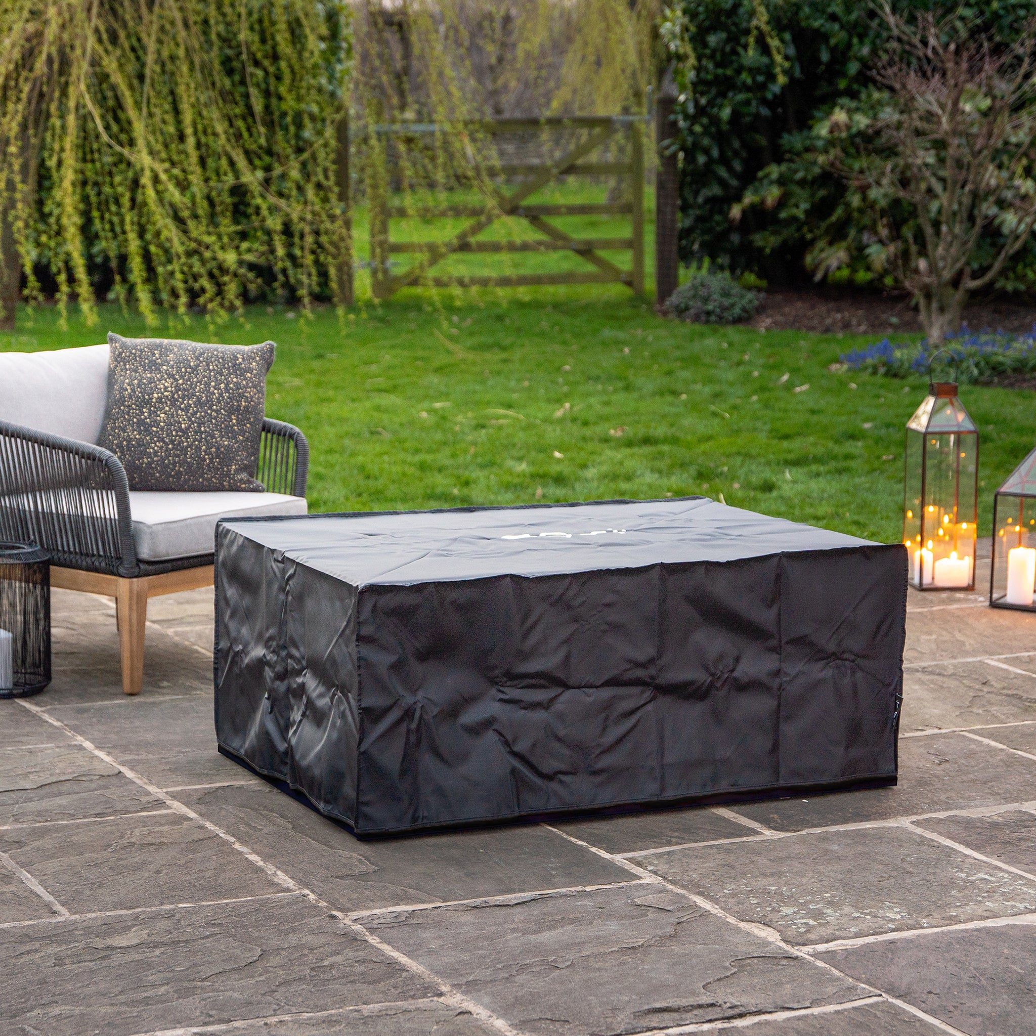An outdoor patio features a weatherproof Cosi Cosiloft 120 Black and Teak Fire Pit Table, elegant wicker chair with cushion, lanterns illuminating the space, and a grassy yard with trees. A tranquil haven crafted from durable teak wood.