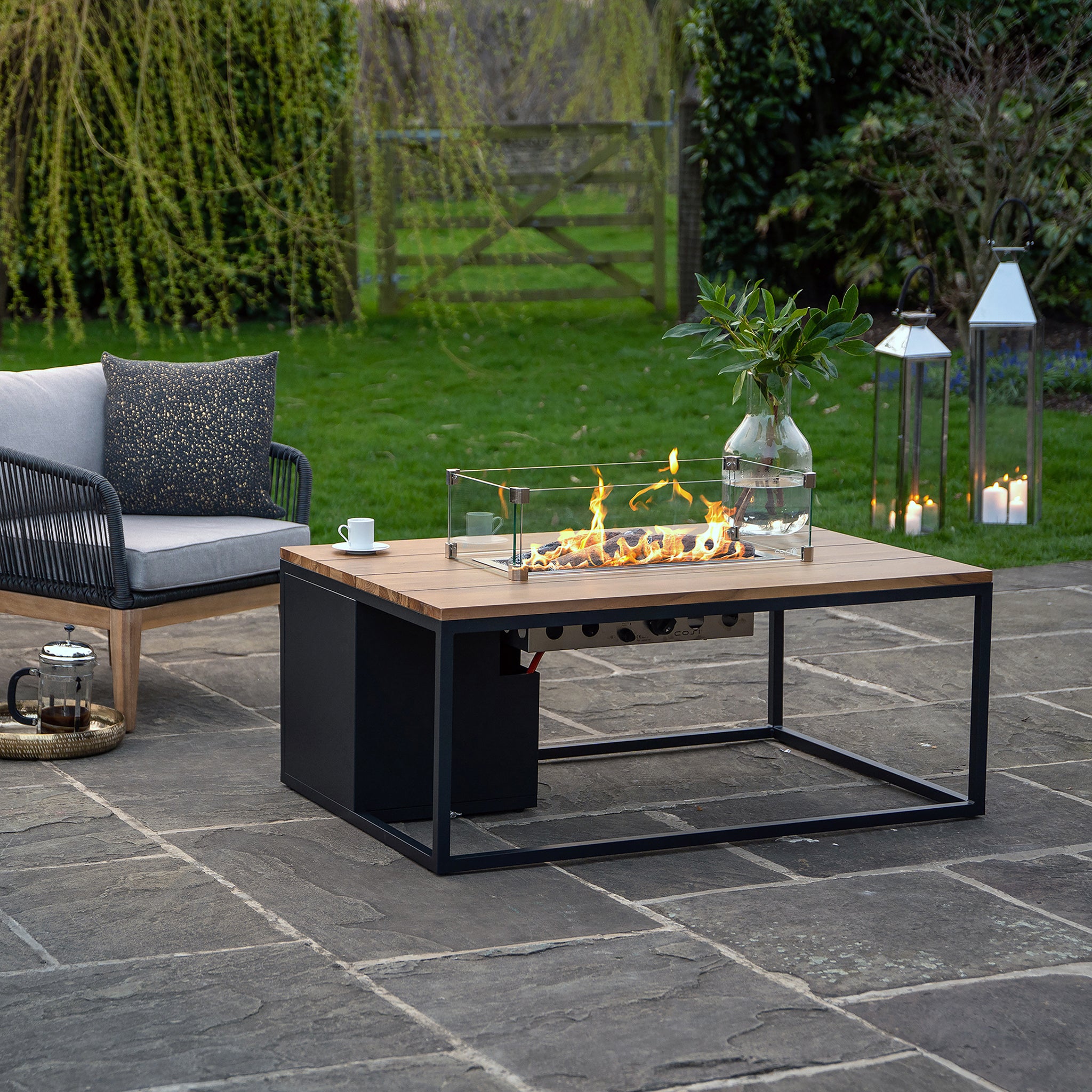 Patio featuring a Cosiloft 120 Black and Teak Fire Pit Table, surrounded by a teak wood sofa, lanterns, and a teapot on stone pavement.