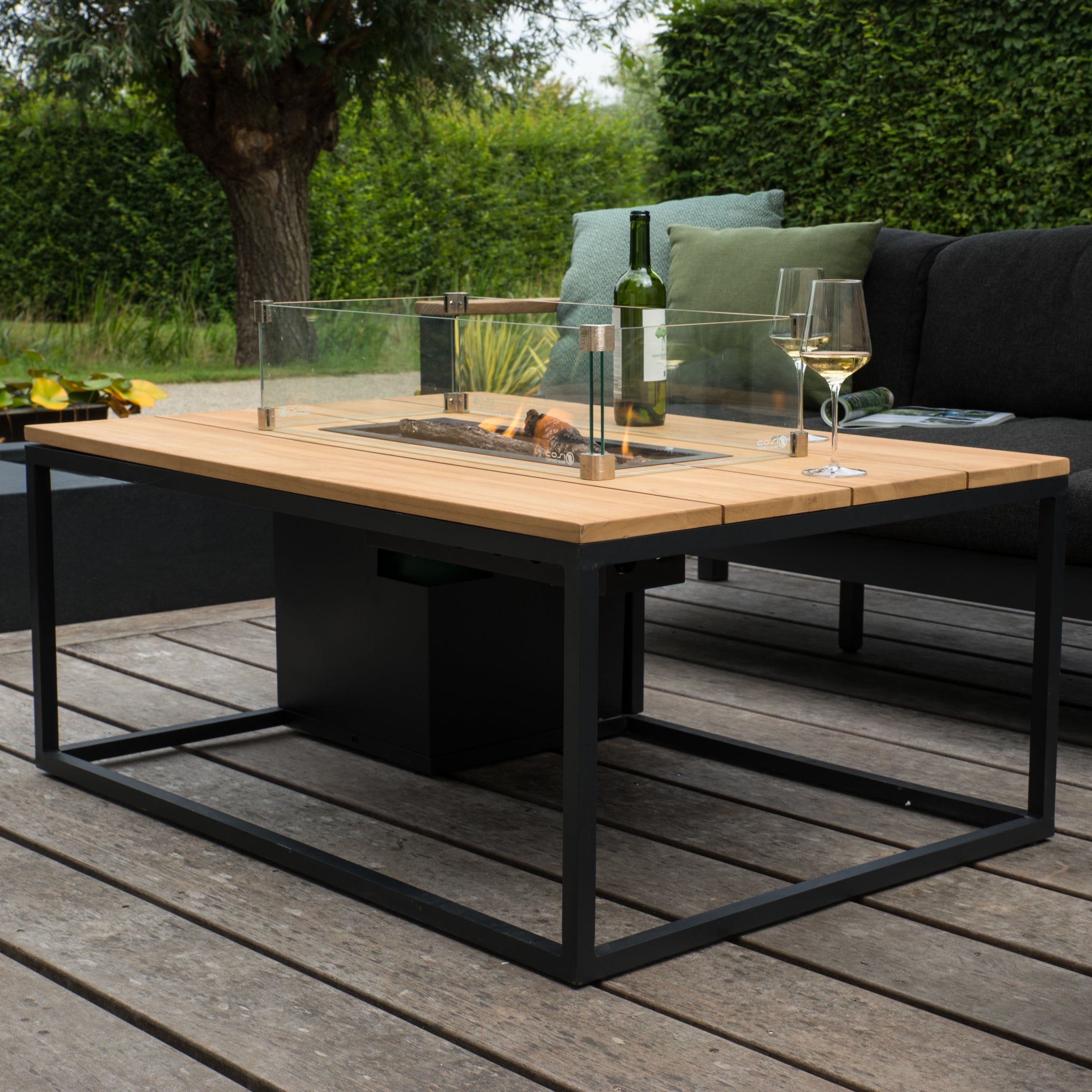 The Cosi - Cosiloft 120 Black and Teak Fire Pit Table sits among cushioned seats on a teak deck, paired with a wine bottle and glass. The weatherproof setup is encircled by lush greenery, ensuring durability and charm.