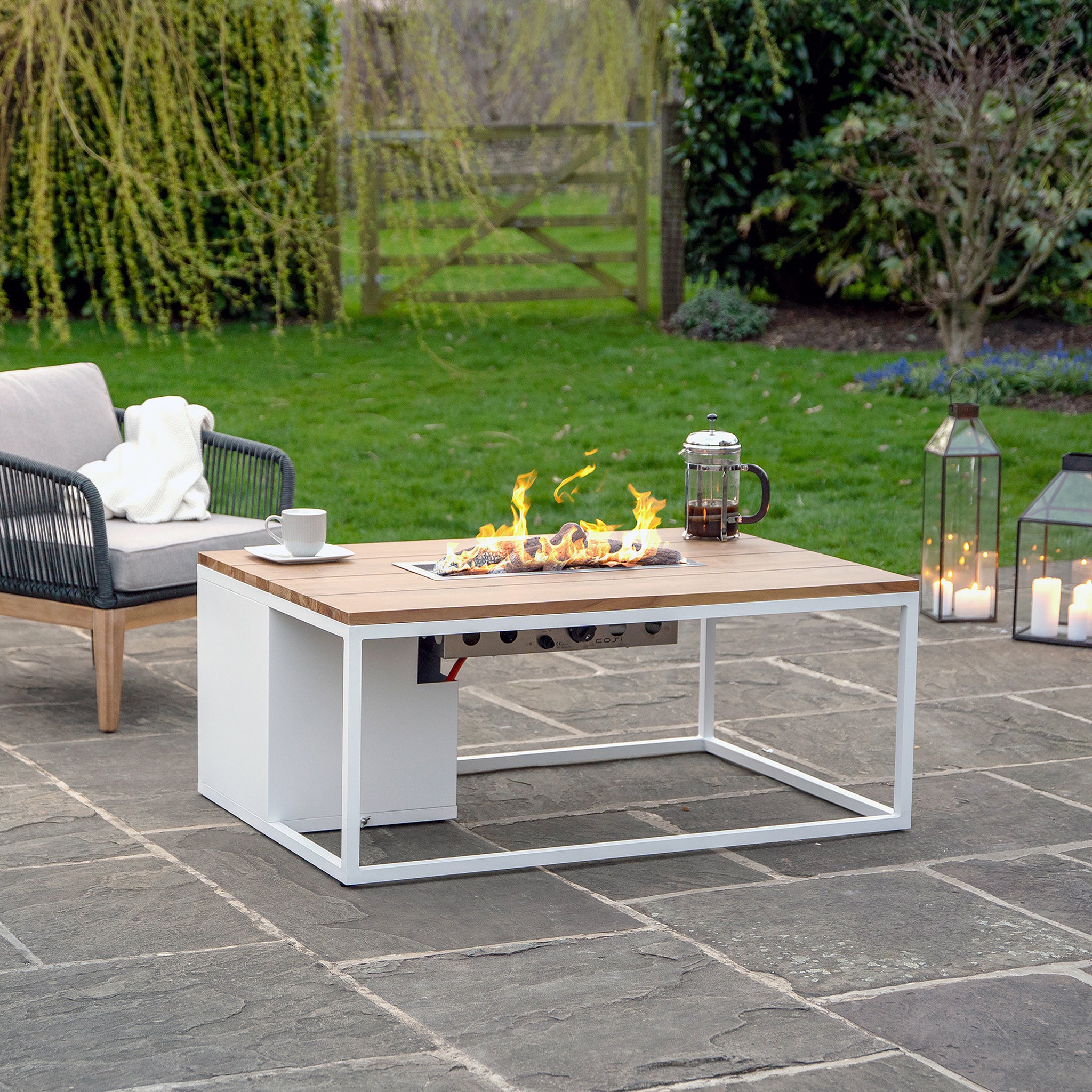 Outdoor patio with a Cosi Cosiloft 120 White and Teak Fire Pit Table, candles, a French press, and a teak wood chair on a stone floor surrounded by greenery.