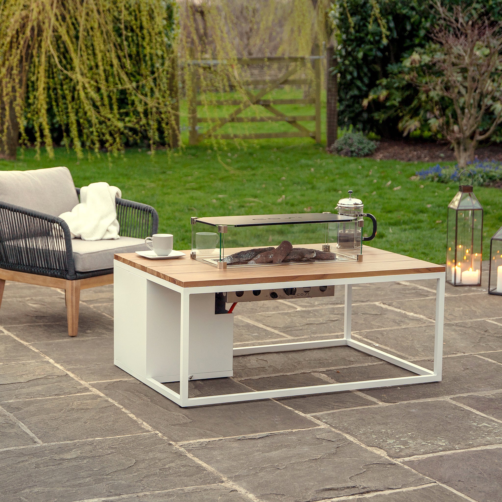 Outdoor lounging is elevated with the Cosi Cosiloft 120 White and Teak Fire Pit Table, paired with a weatherproof chair and chic lantern on stone tiles, surrounded by lush greenery.