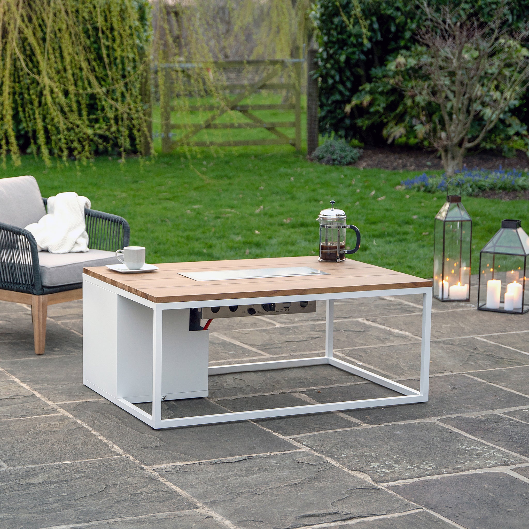 The outdoor patio showcases a Cosi - Cosiloft 120 White and Teak Fire Pit Table, a French press, and a teak wood chair for ideal lounging. Weatherproof lanterns illuminate the stone surface with a warm glow, enhancing the inviting ambiance.