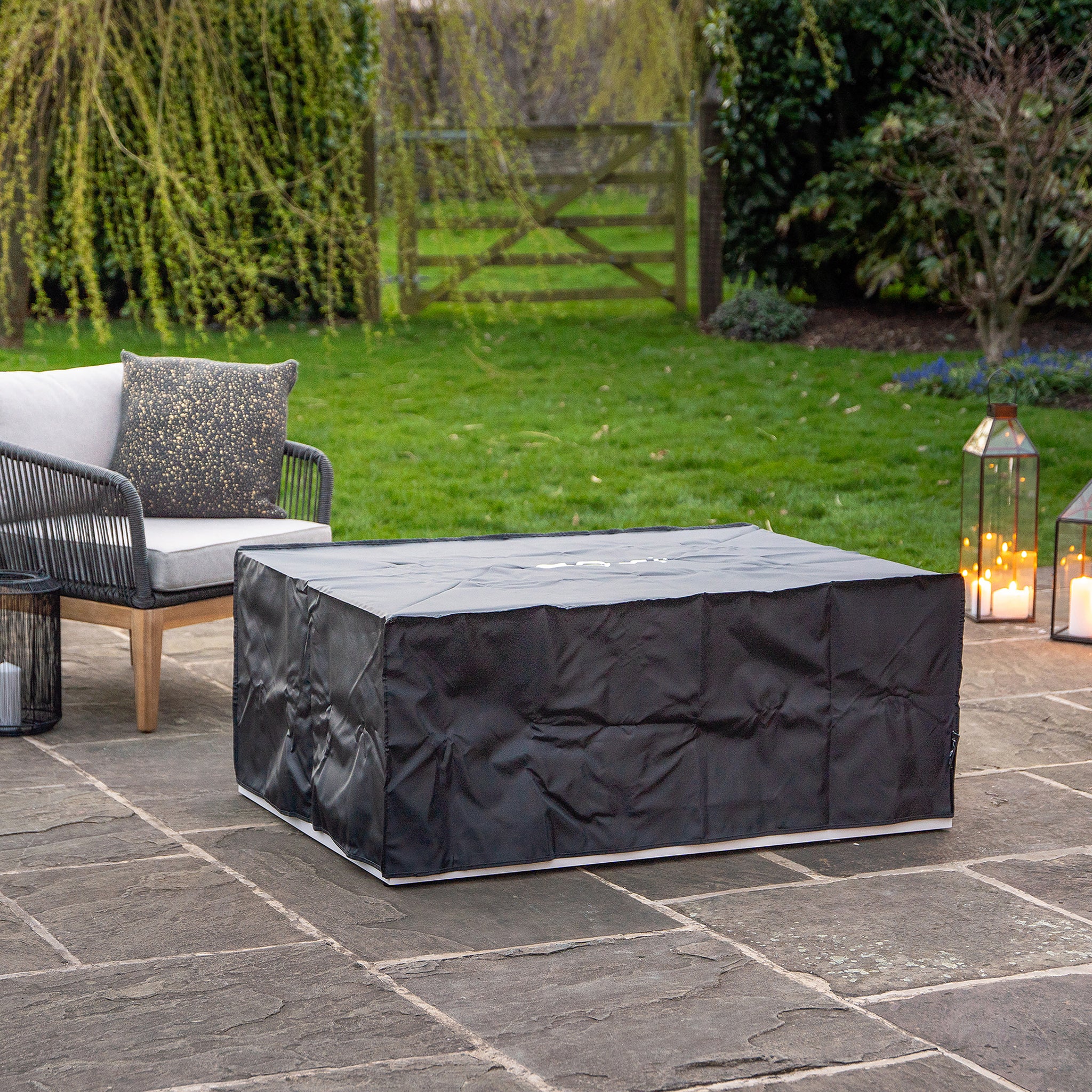 Outdoor patio scene featuring a weatherproof Cosi - Cosiloft 120 White and Teak Fire Pit Table, wicker sofa, and lanterns amidst lush greenery, perfect for outdoor lounging.
