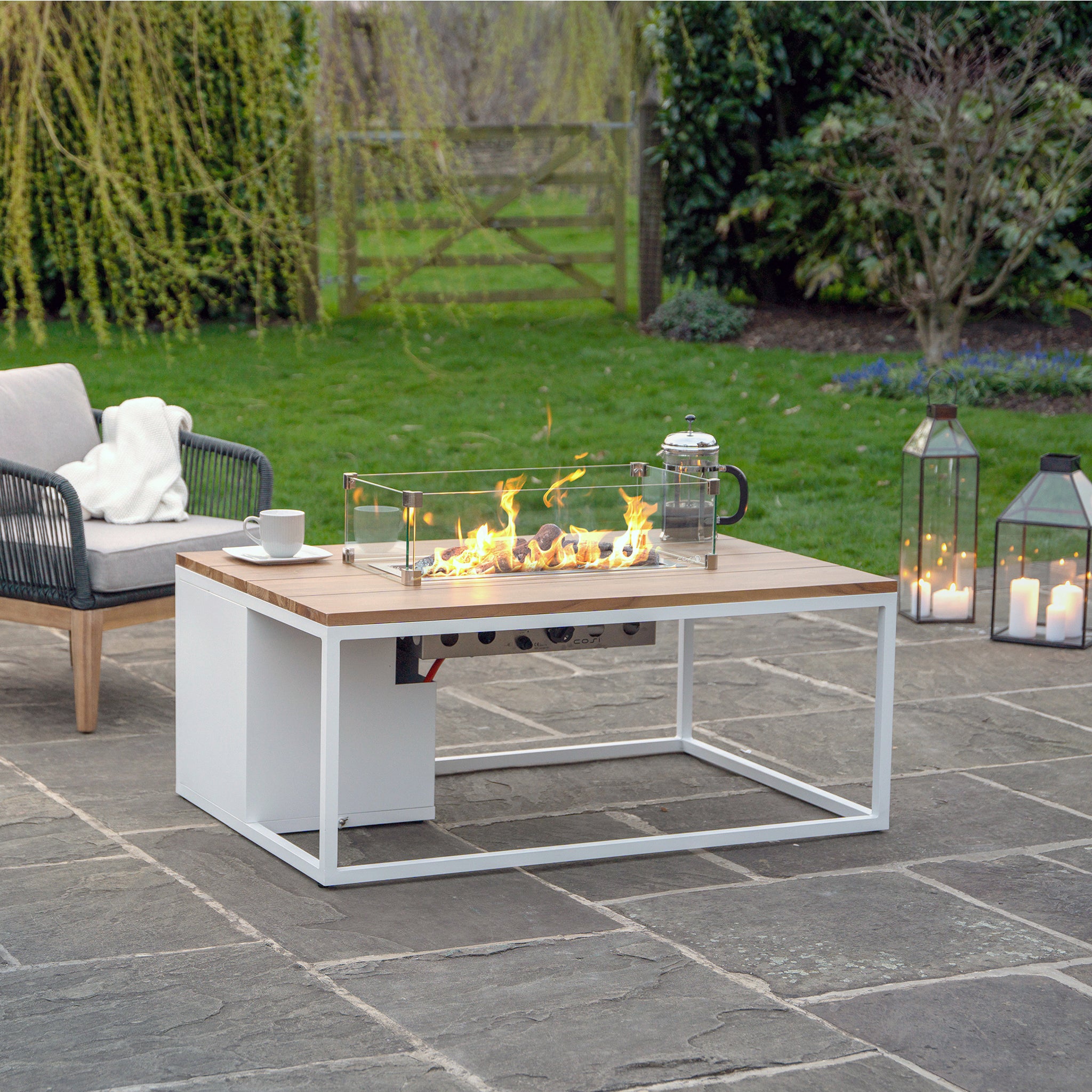The Cosi - Cosiloft 120 White and Teak Fire Pit Table enhances a stone patio with its weatherproof lounging appeal, complemented by durable teak wood seating amidst greenery and lanterns.