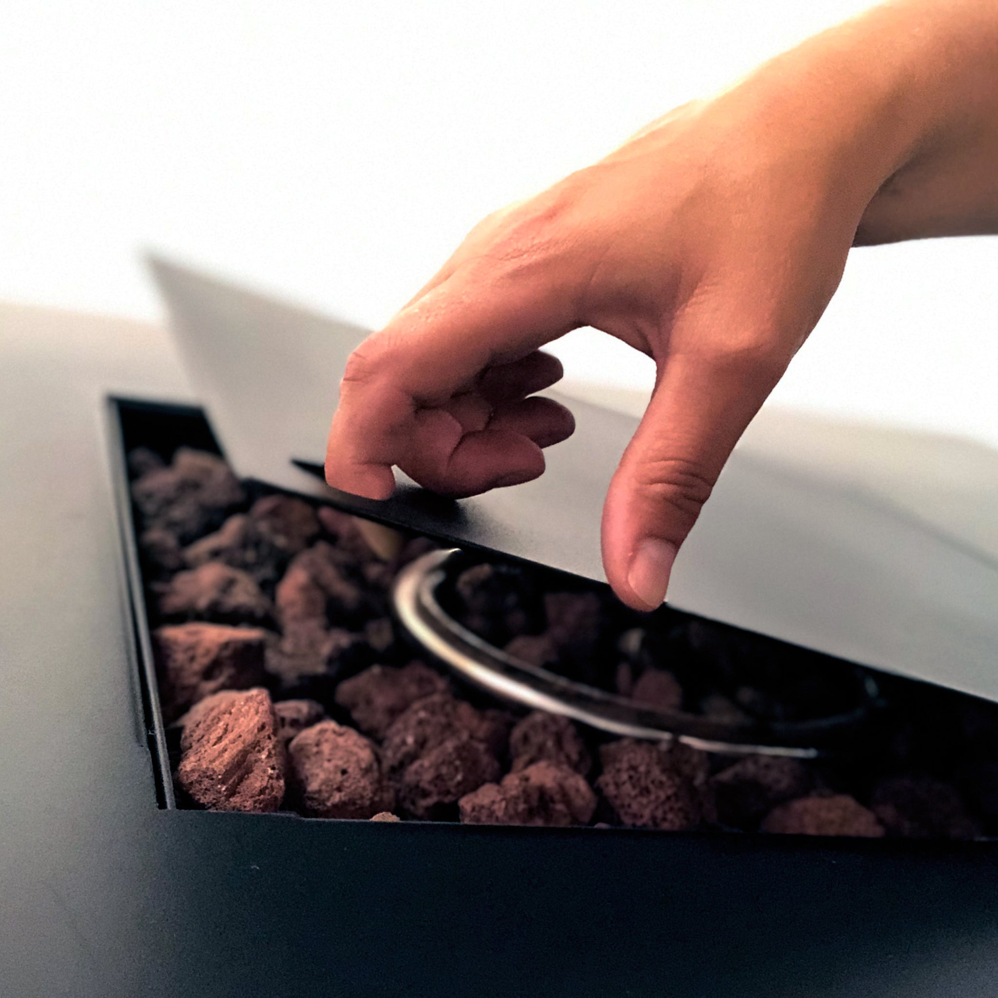 A hand lifts the metal lid of the Cosi - Cosibrixx 90 Anthracite Fire Pit Table, unveiling glowing lava rocks and a sturdy metal coil beneath its anthracite exterior.