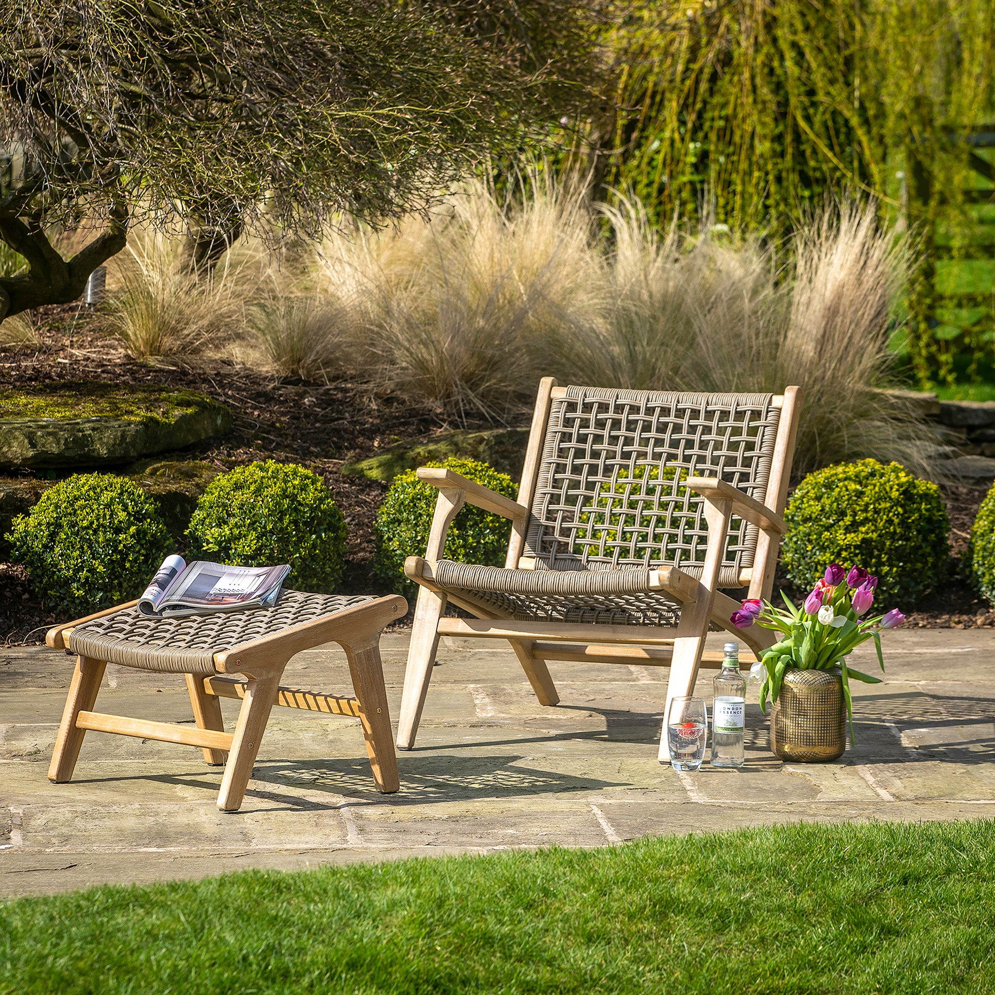 The Pacific Lifestyle - Sesto Lounge Chair & Hocker Set elegantly graces the patio, accompanied by a magazine, tulips in a vase, and refreshing drinks in a serene garden. This setup invites relaxation while showcasing quality garden furniture with its durable waterproof seat against nature's elements.