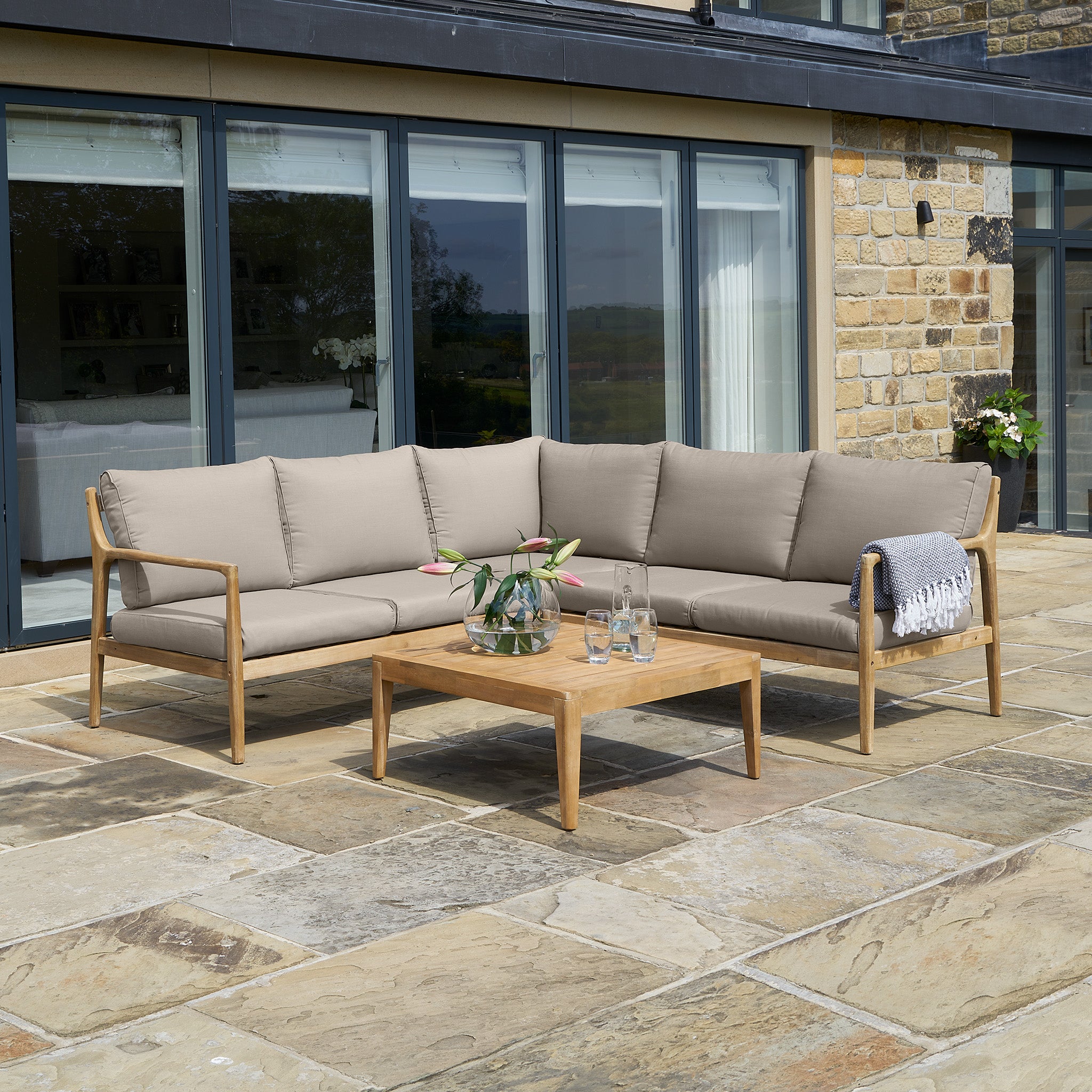 A cozy patio showcases the Pacific Lifestyle Malta Lounge Corner Set in Natural Taupe, paired with a wooden coffee table and glass vase. A gray blanket elegantly drapes over the acacia wood frame, all on stone tiles near welcoming glass doors.