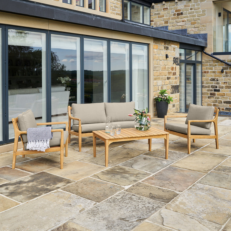 A modern patio featuring the Pacific Lifestyle - Malta Lounge Seating Set in Royal Taupe with weatherproof furniture, complemented by a sleek coffee table. The Acacia wood frame pairs elegantly with glass doors and stone paving, creating an inviting outdoor oasis.