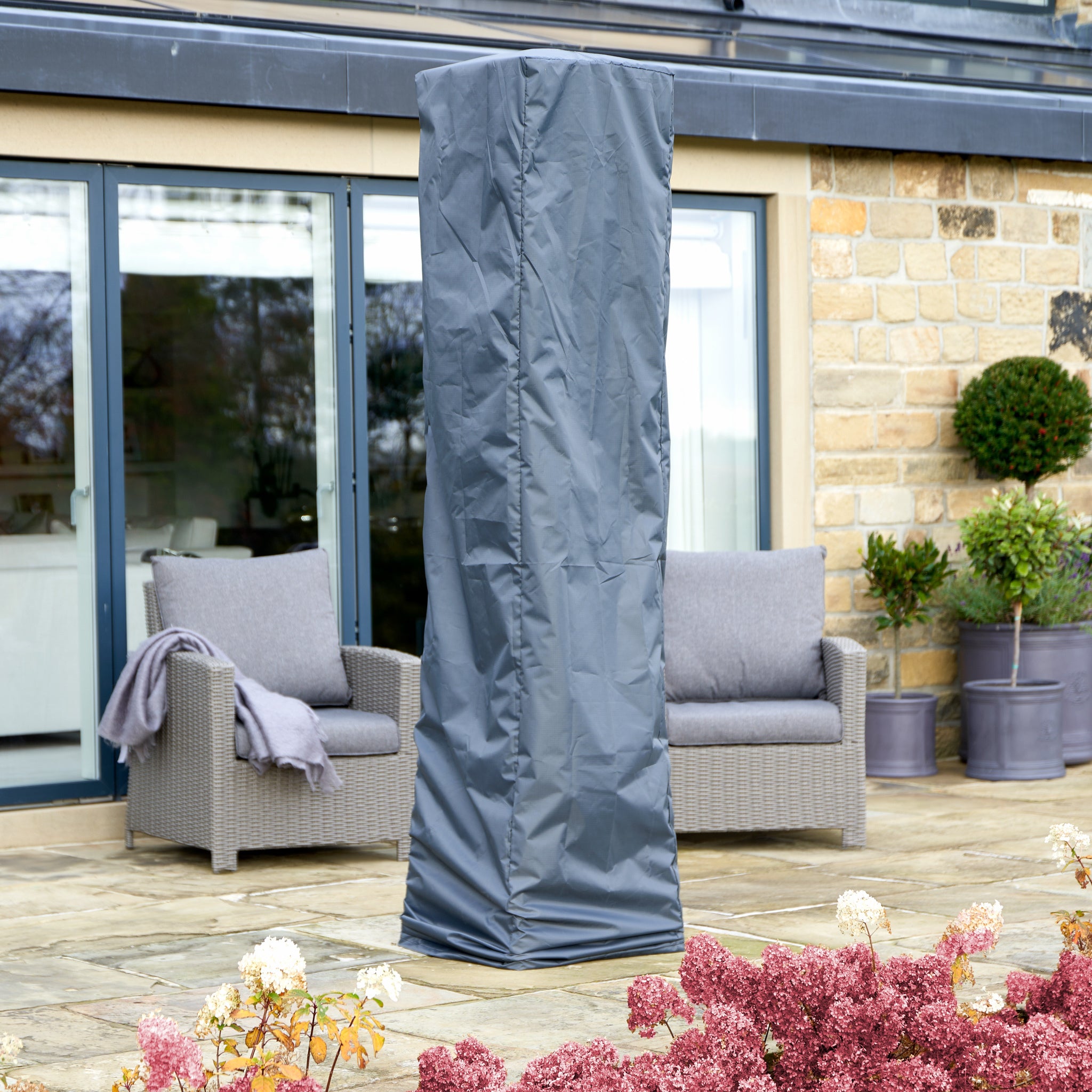 The Pacific Lifestyle Quadrilateral Patio Heater Cover in tall grey with Aerocover protection keeps your patio heating setup pristine, surrounded by outdoor furniture and potted plants.